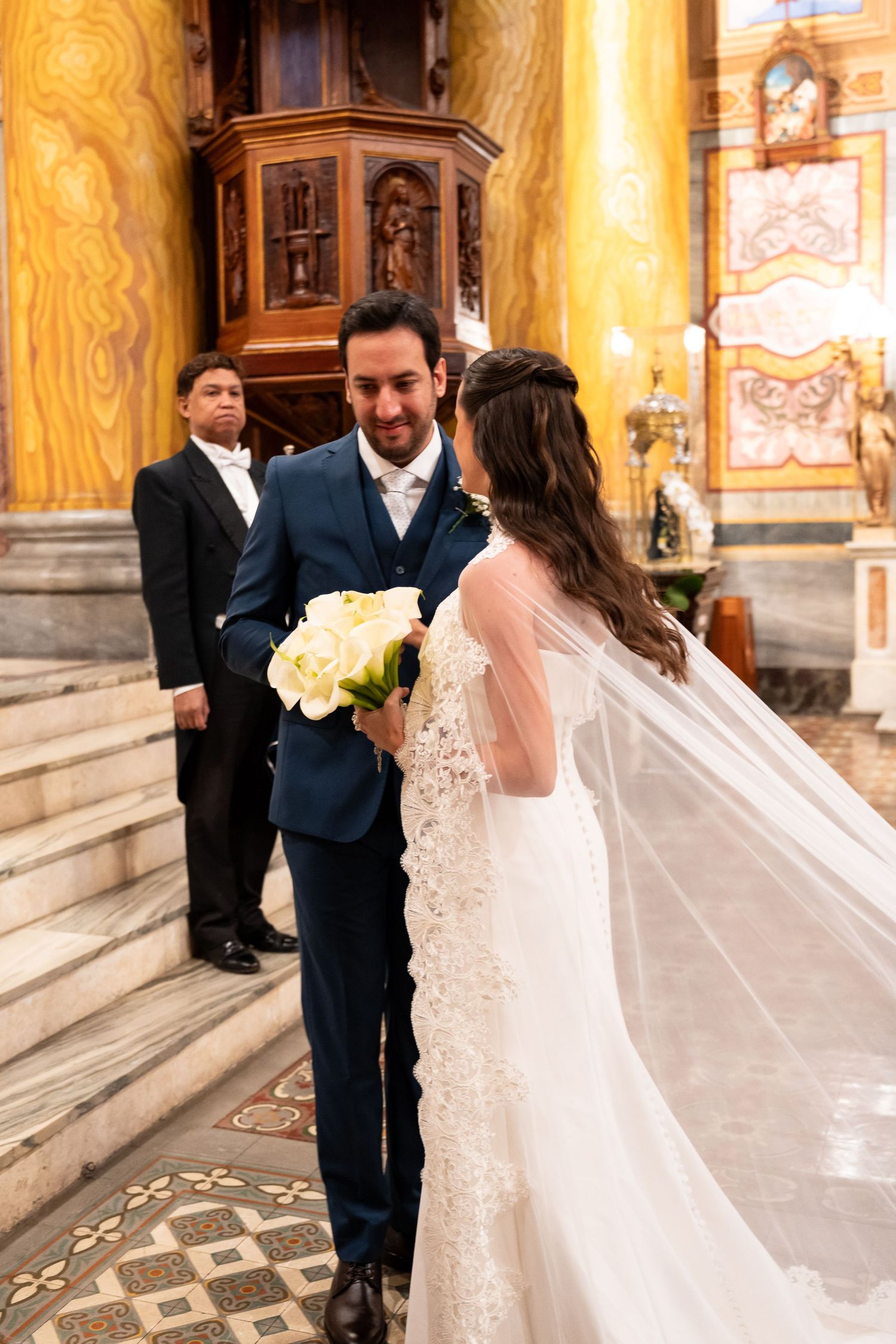 Bride and groom share a tender moment at church altar, bride holding white roses bouquet, wearing lace veil.