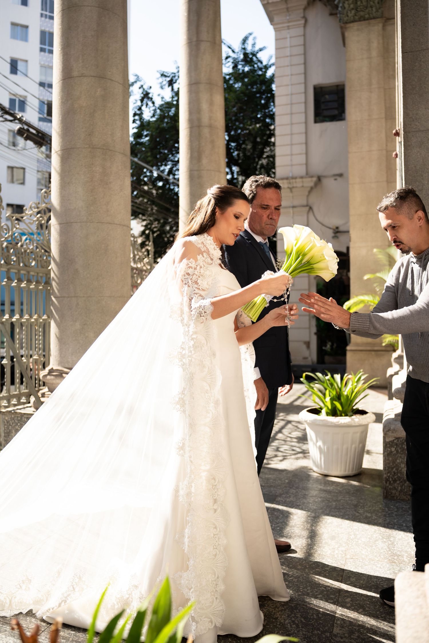 Bride in elegant lace wedding gown receiving white calla lily bouquet from groomsman outside a church.