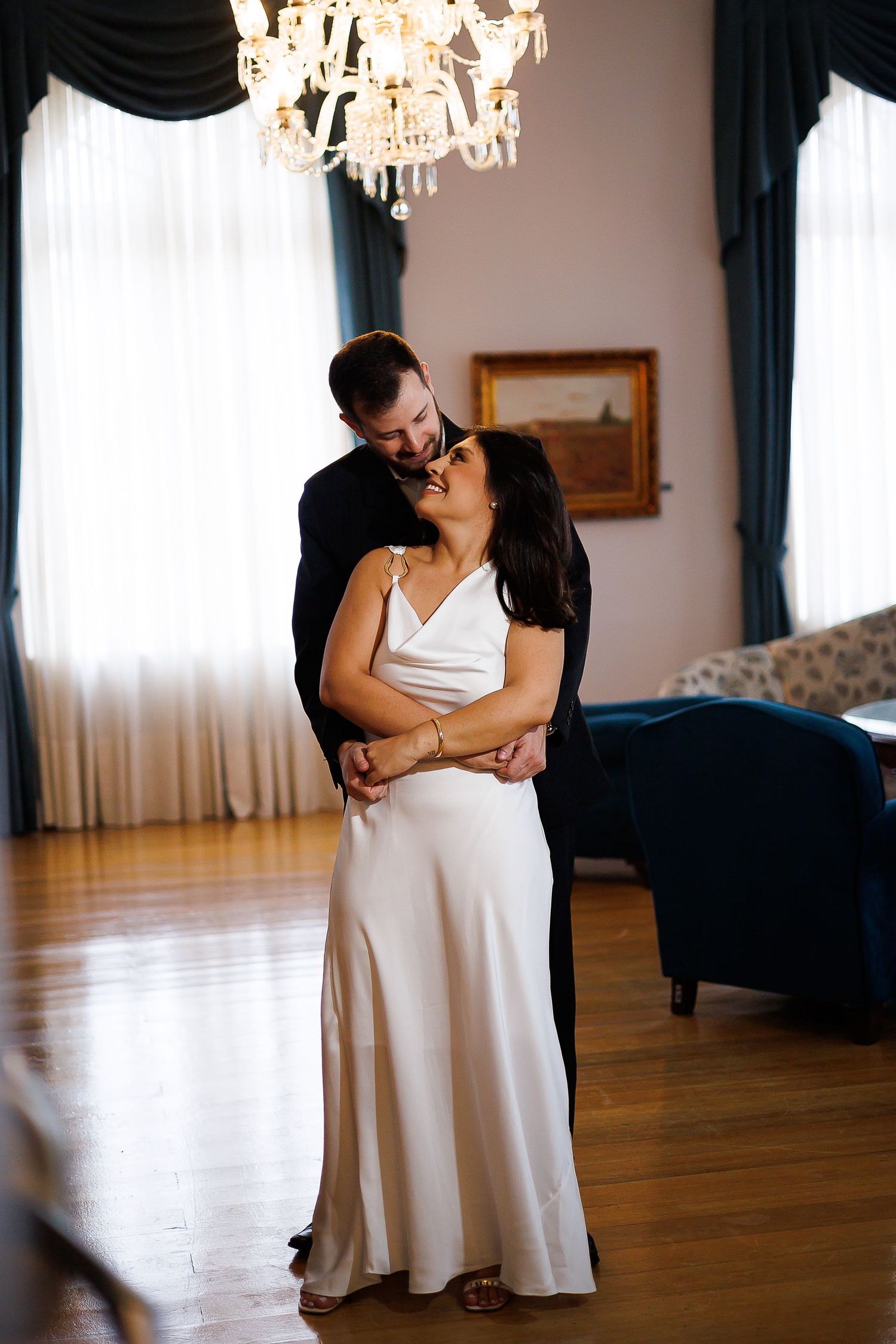 Bride and groom share a romantic embrace in an elegant room with hardwood floors and a crystal chandelier.
