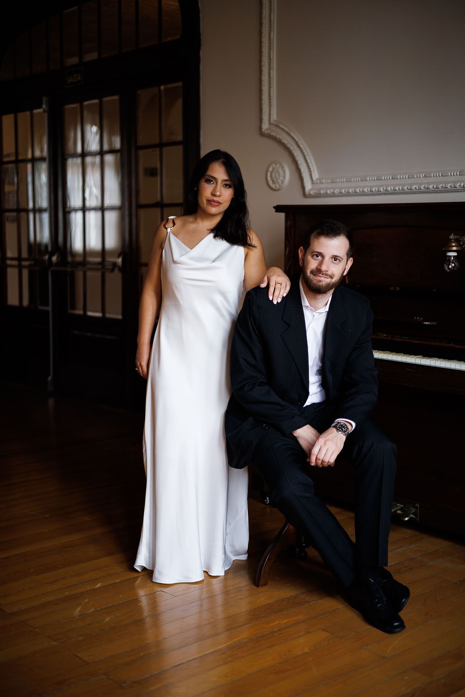 Elegant couple posing near a piano, woman in white gown and man in black suit, in a classic interior setting.