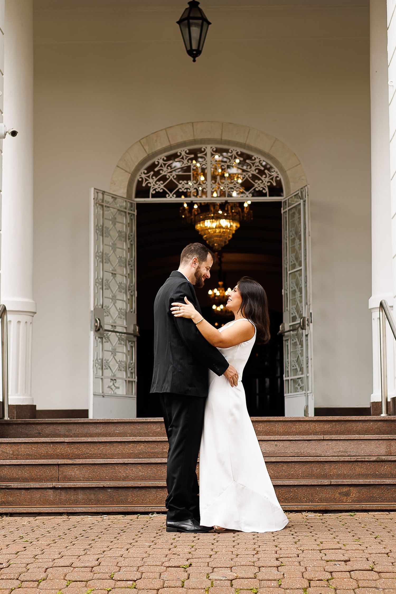 Bride and groom embrace on brick steps before elegant white venue with chandelier and arched doorway.
