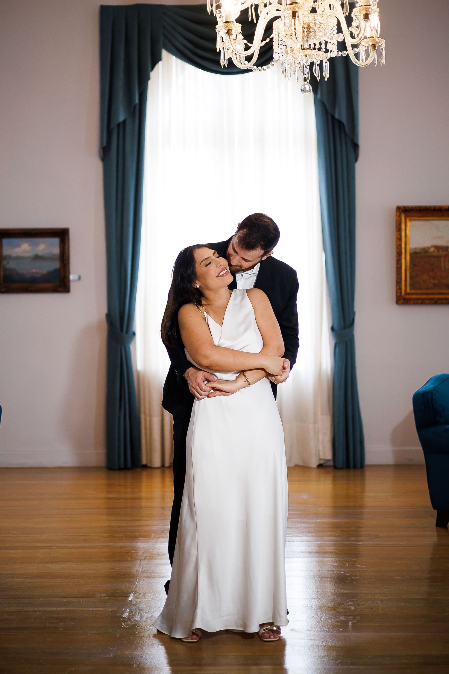 A bride and groom share a tender embrace in an elegant room with chandelier, hardwood floors, and teal drapes.