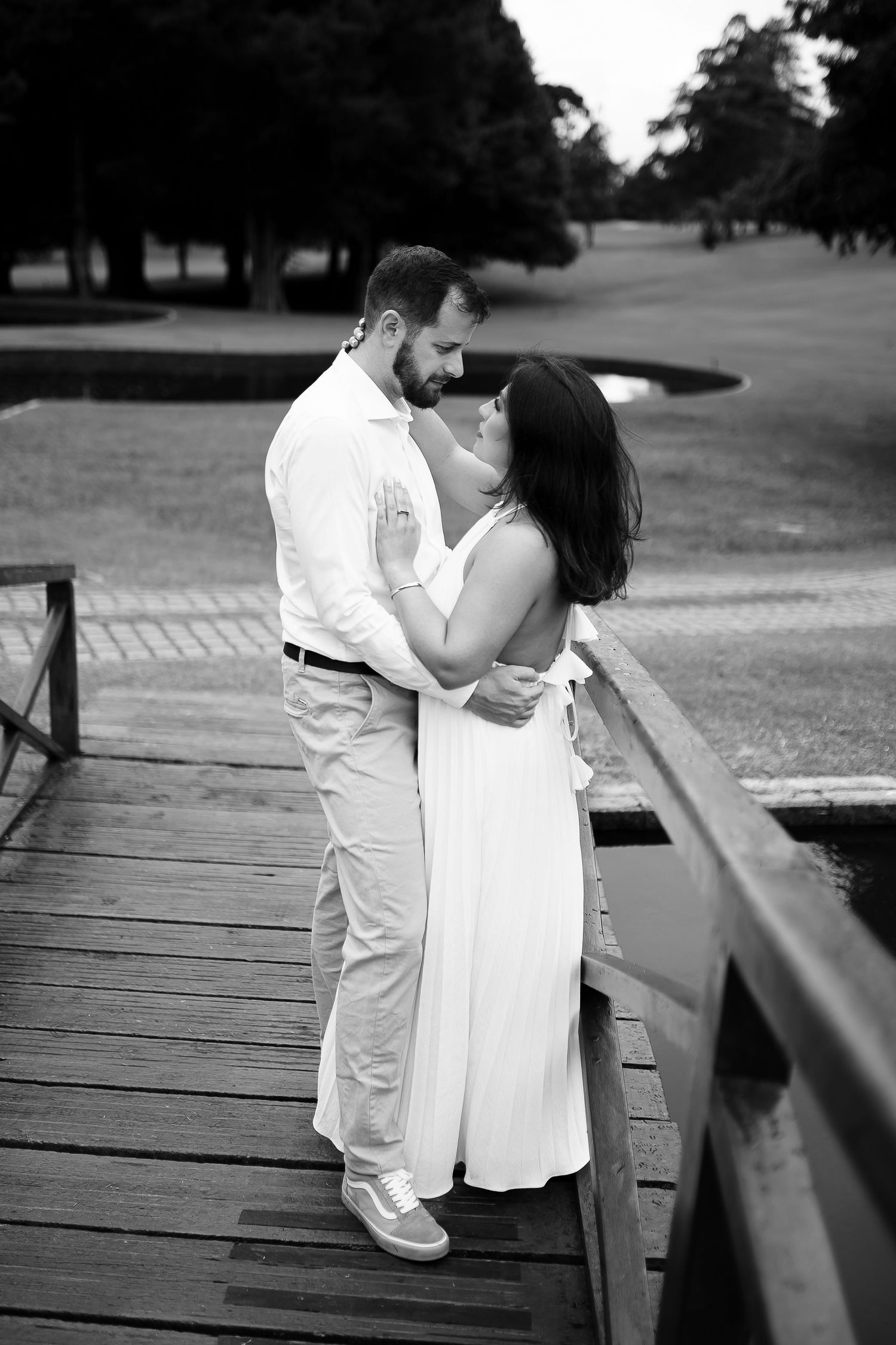 Black and white photo of a couple embracing romantically on a wooden bridge surrounded by lush greenery.