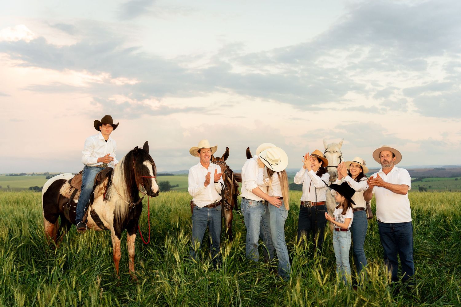 Family in cowboy hats posing with horses in a green field at sunset, celebrating a country lifestyle.