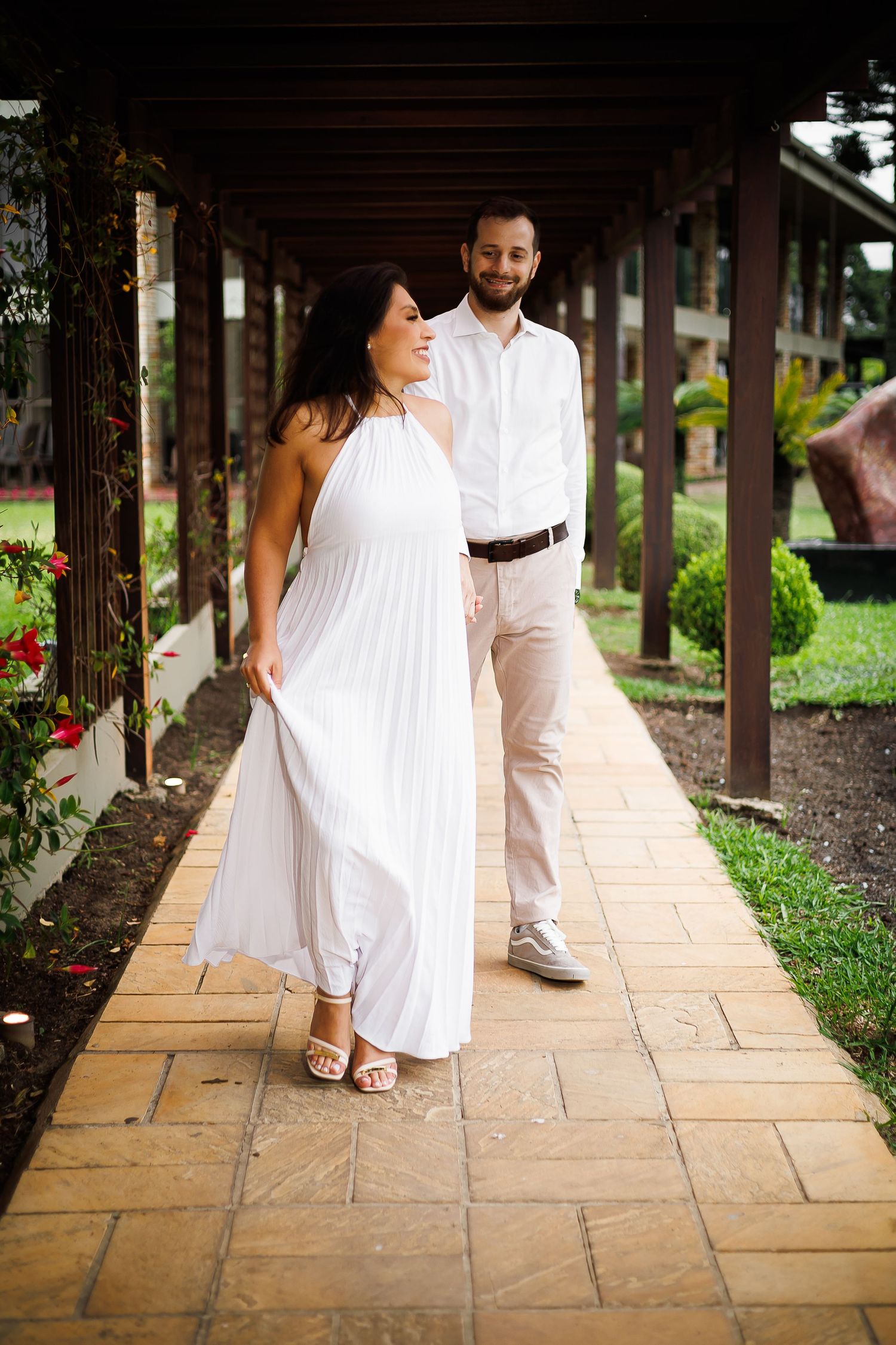 A couple walks hand-in-hand under a wooden pergola, woman in white dress, man in white shirt and beige pants.