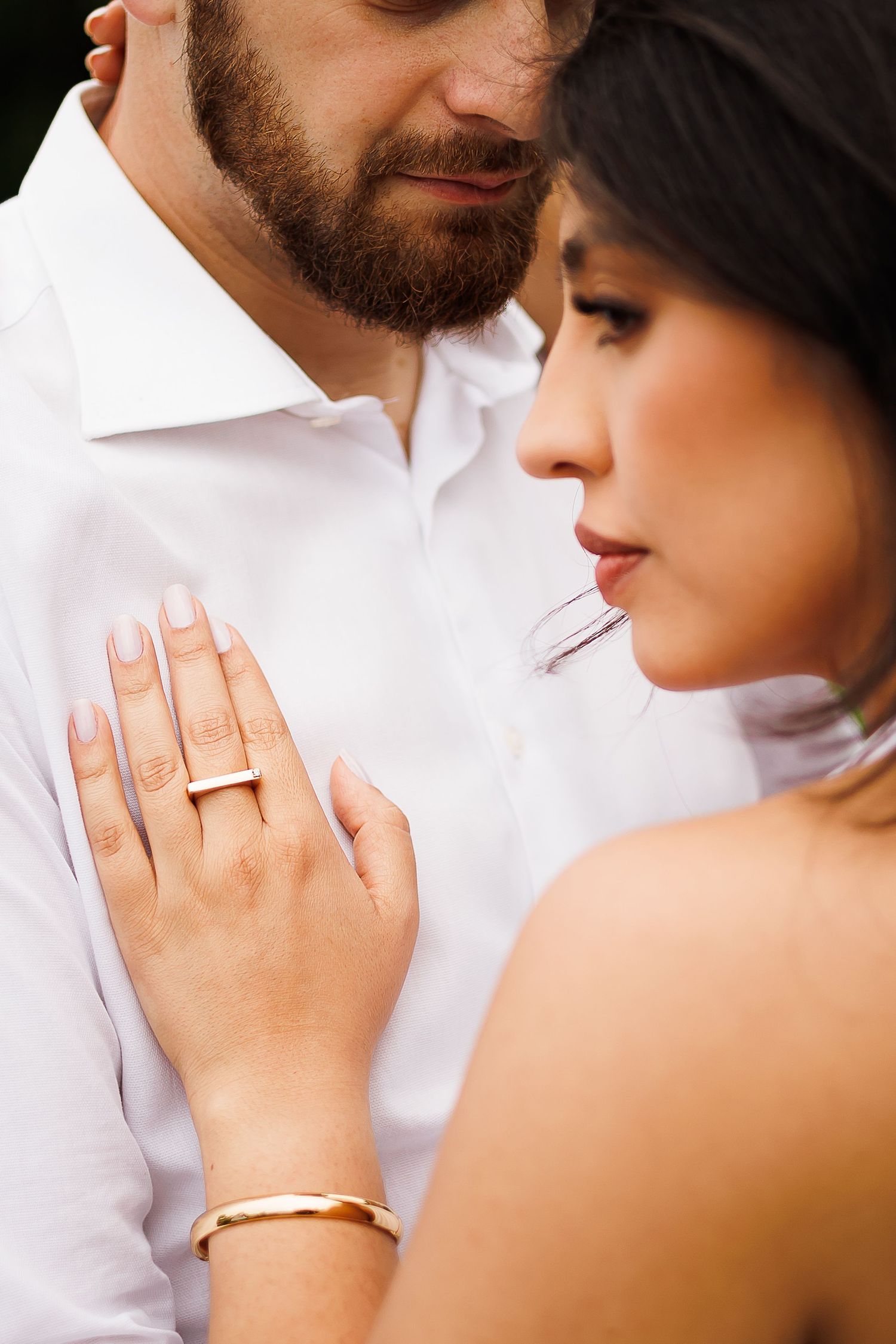 Couple in romantic pose, woman wearing gold ring and bracelet rests hand on mans white shirt chest.