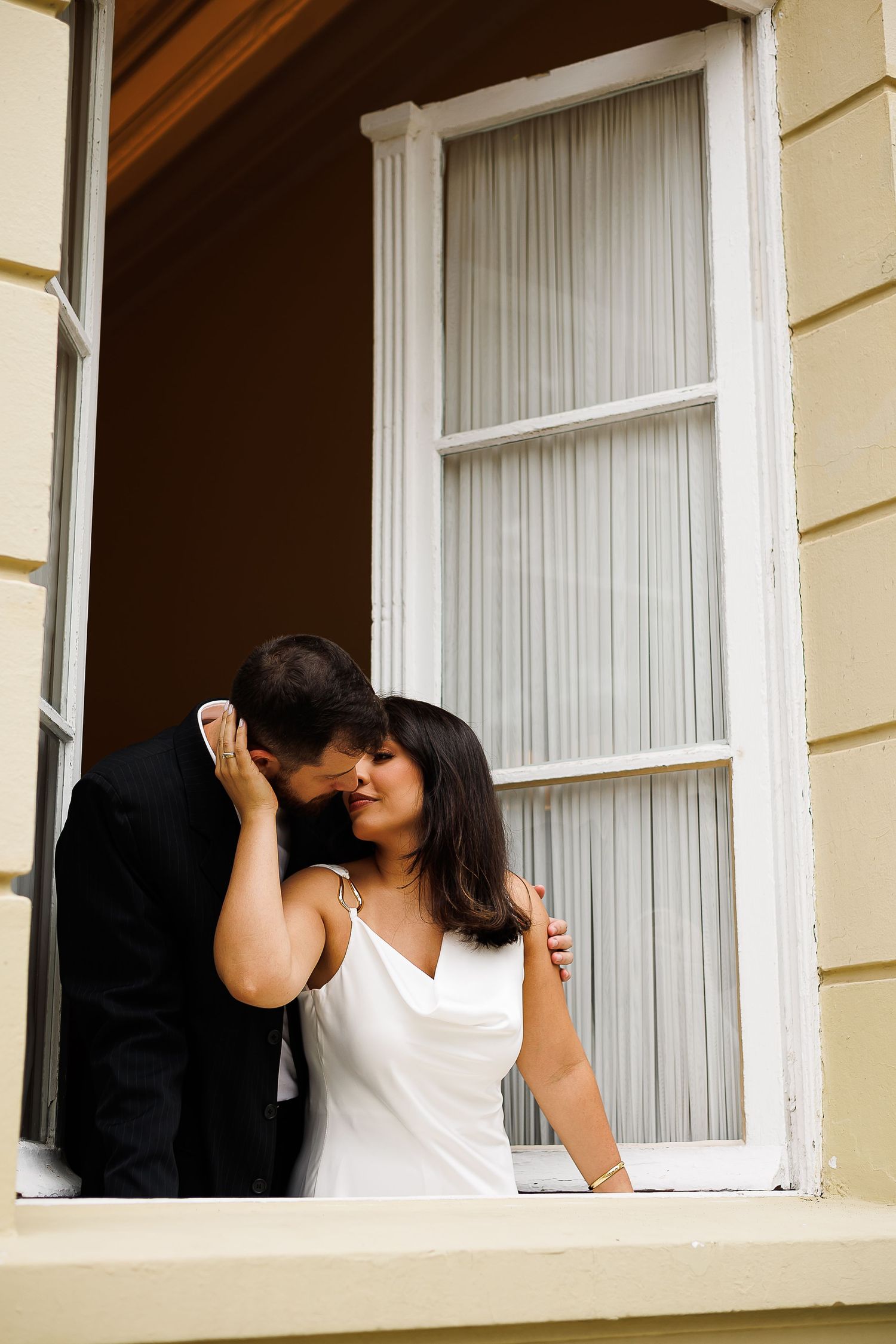 Romantic couple sharing a kiss by an open window, woman in white dress and man in black suit.