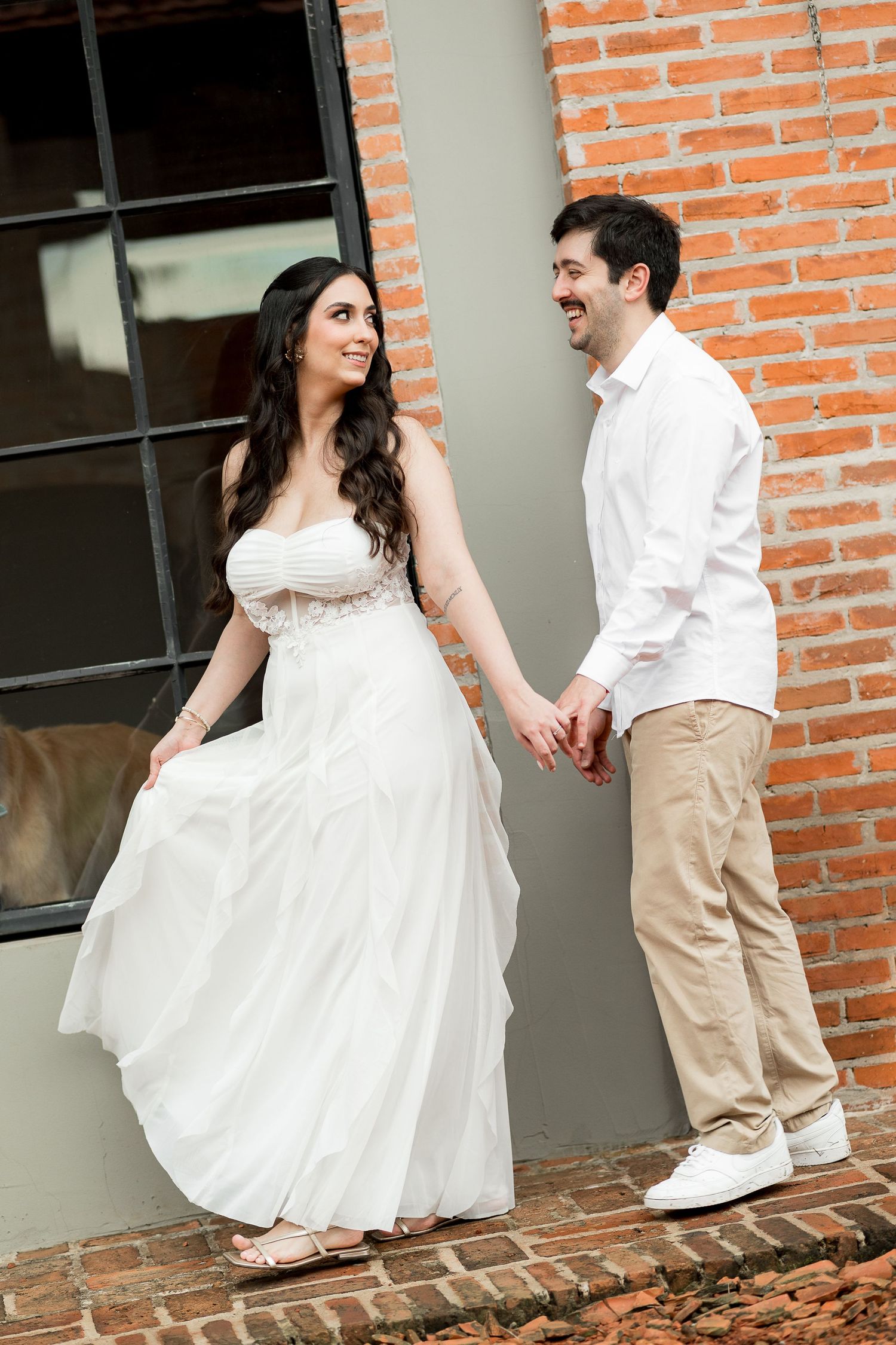 A smiling couple holds hands outdoors, the woman in a flowing white wedding gown and the man in a white shirt and khakis.