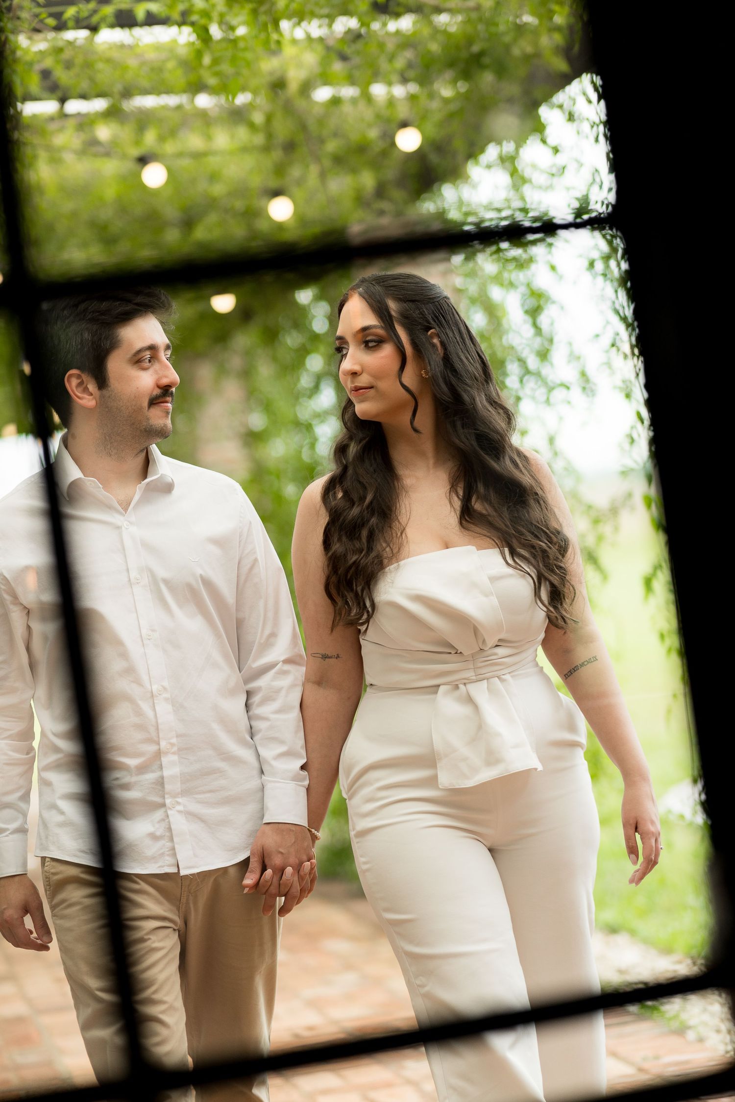 Couple in white outfits holding hands under string lights and green pergola in romantic outdoor setting.