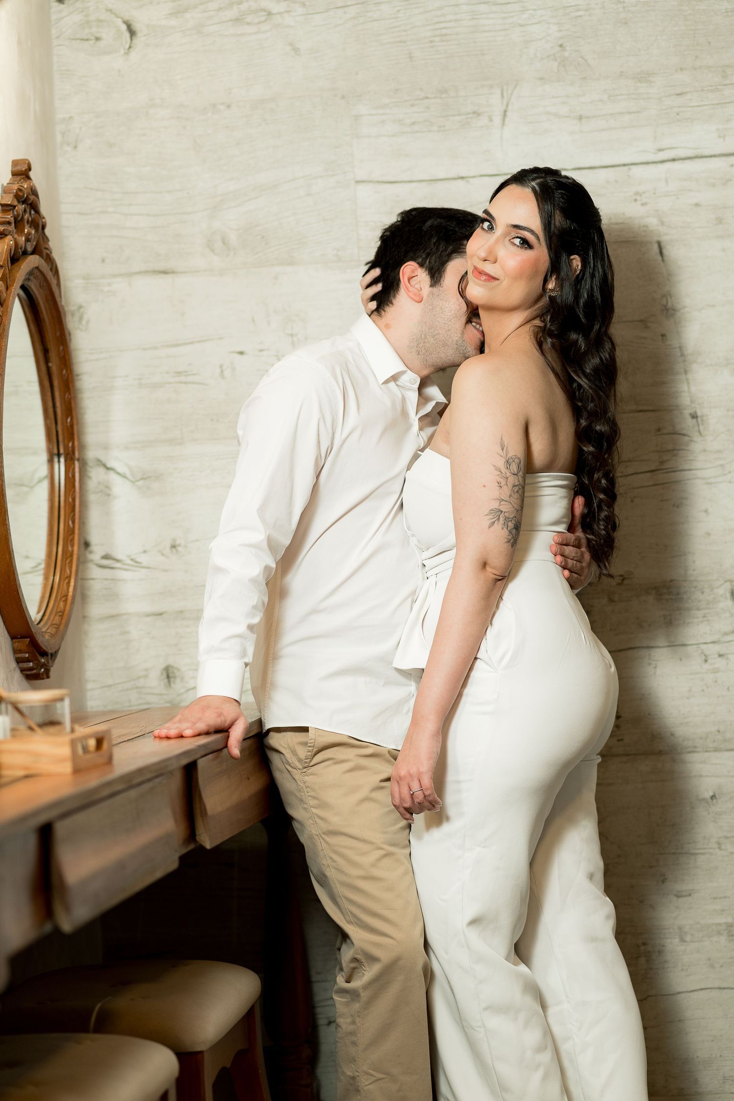 Romantic couple in white outfits posing near vintage mirror, man kissing woman's cheek in elegant rustic interior.