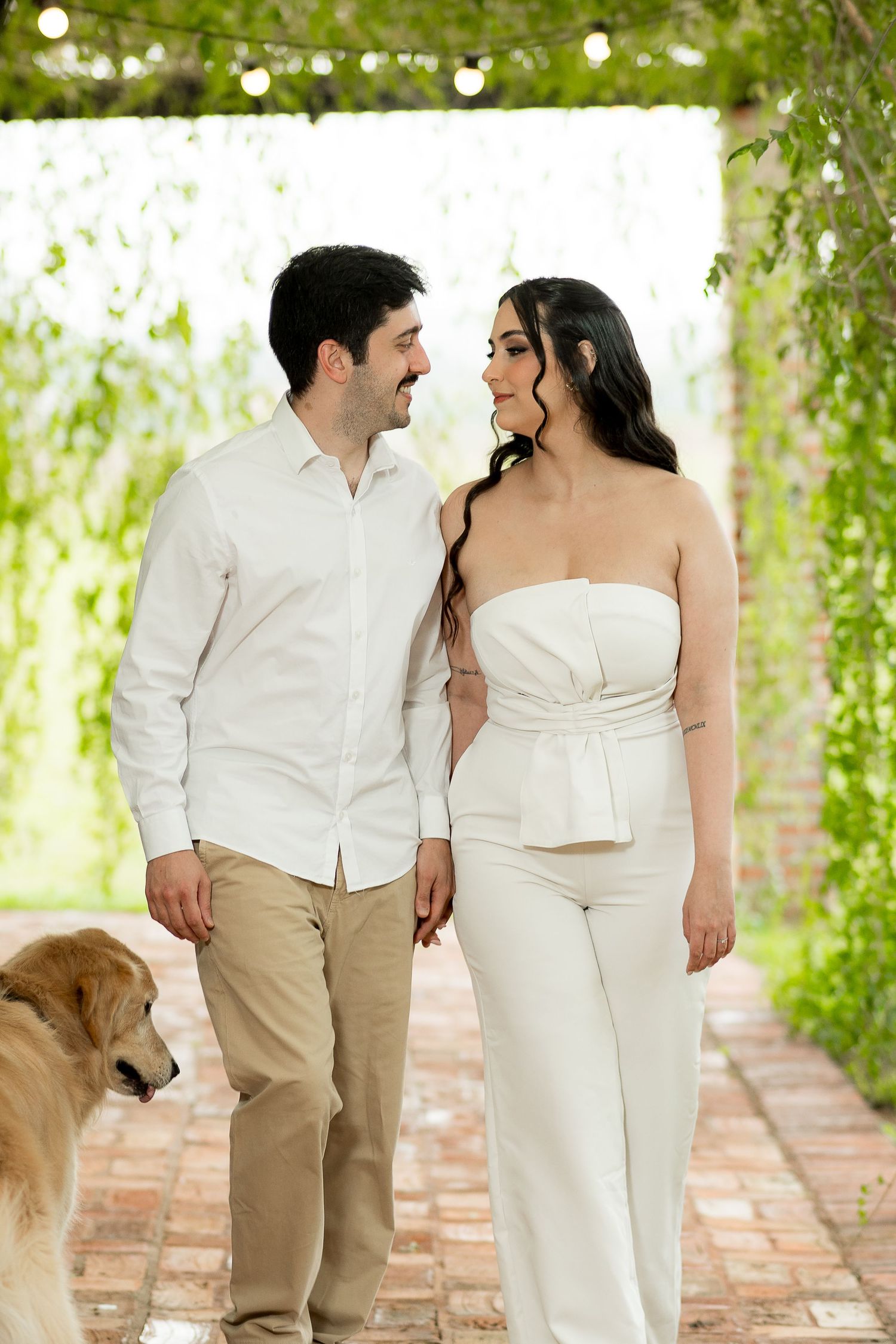 Couple in matching white outfits walking together under a green pergola with a golden retriever on a brick path.
