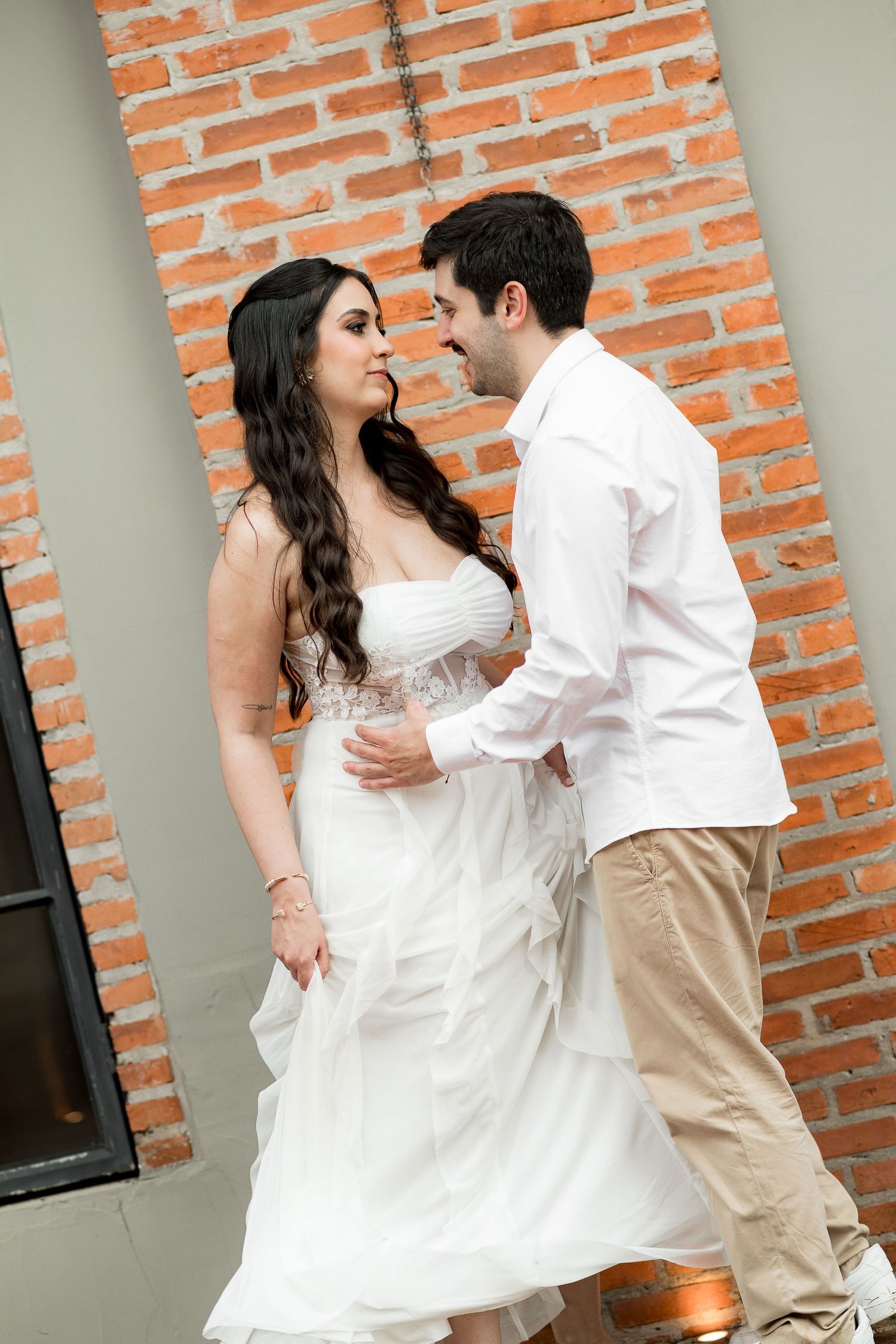 Couple in wedding attire dancing romantically in front of a brick wall, bride in white gown, groom in white shirt.