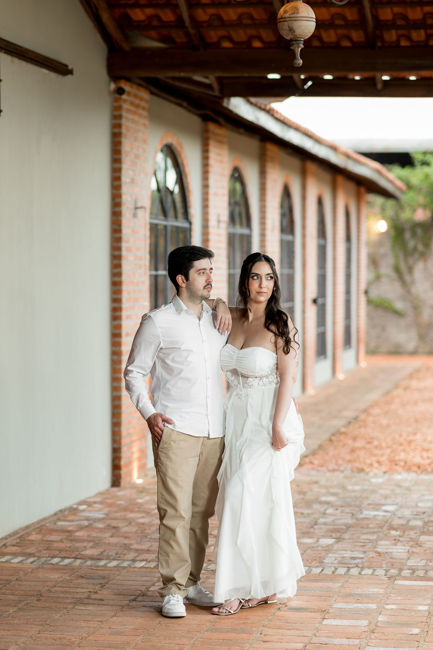 Elegant couple posing in white wedding attire along a rustic brick colonnade walkway.