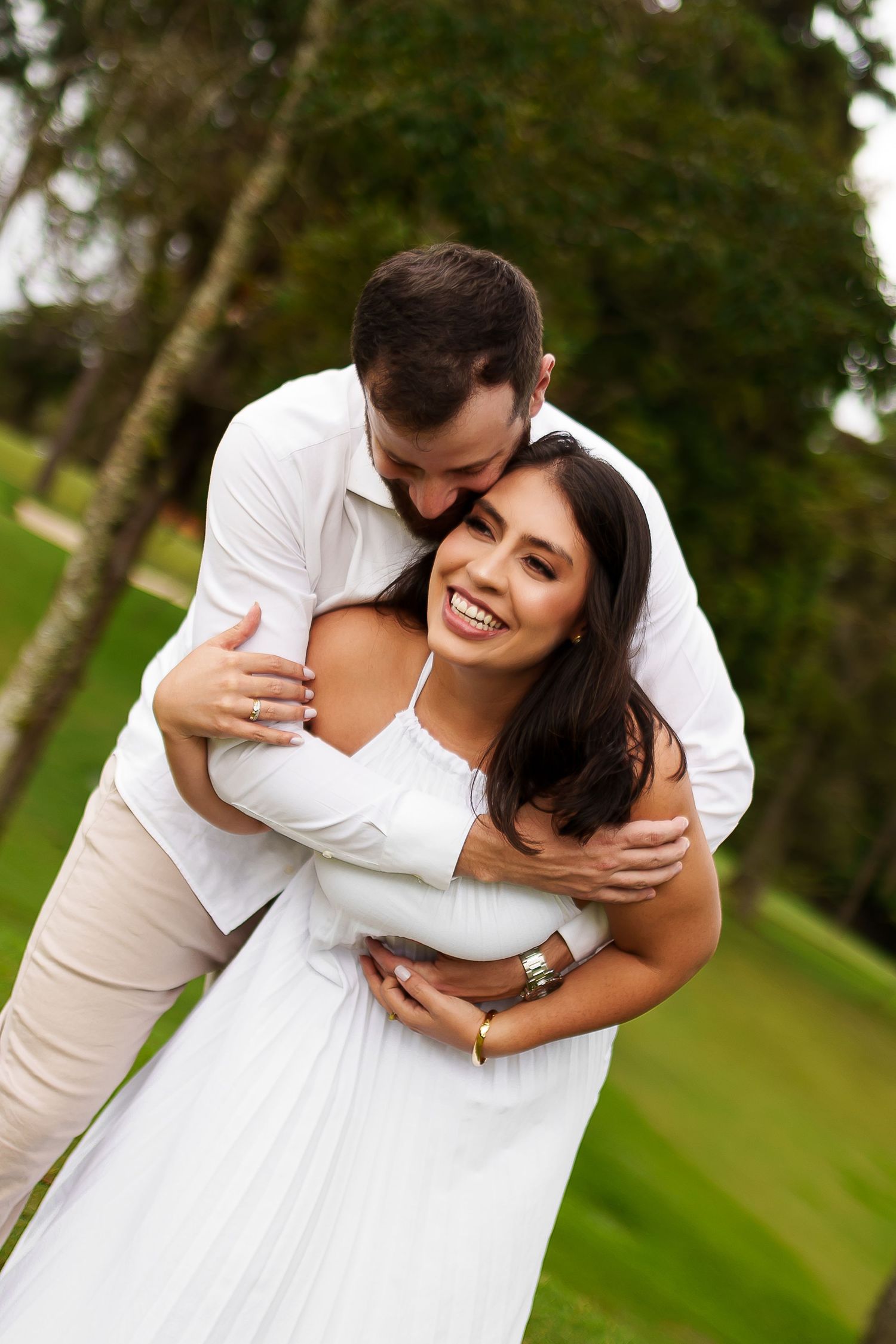 A smiling couple in white outfits embrace outdoors, the man hugging the woman from behind on a lush green lawn.