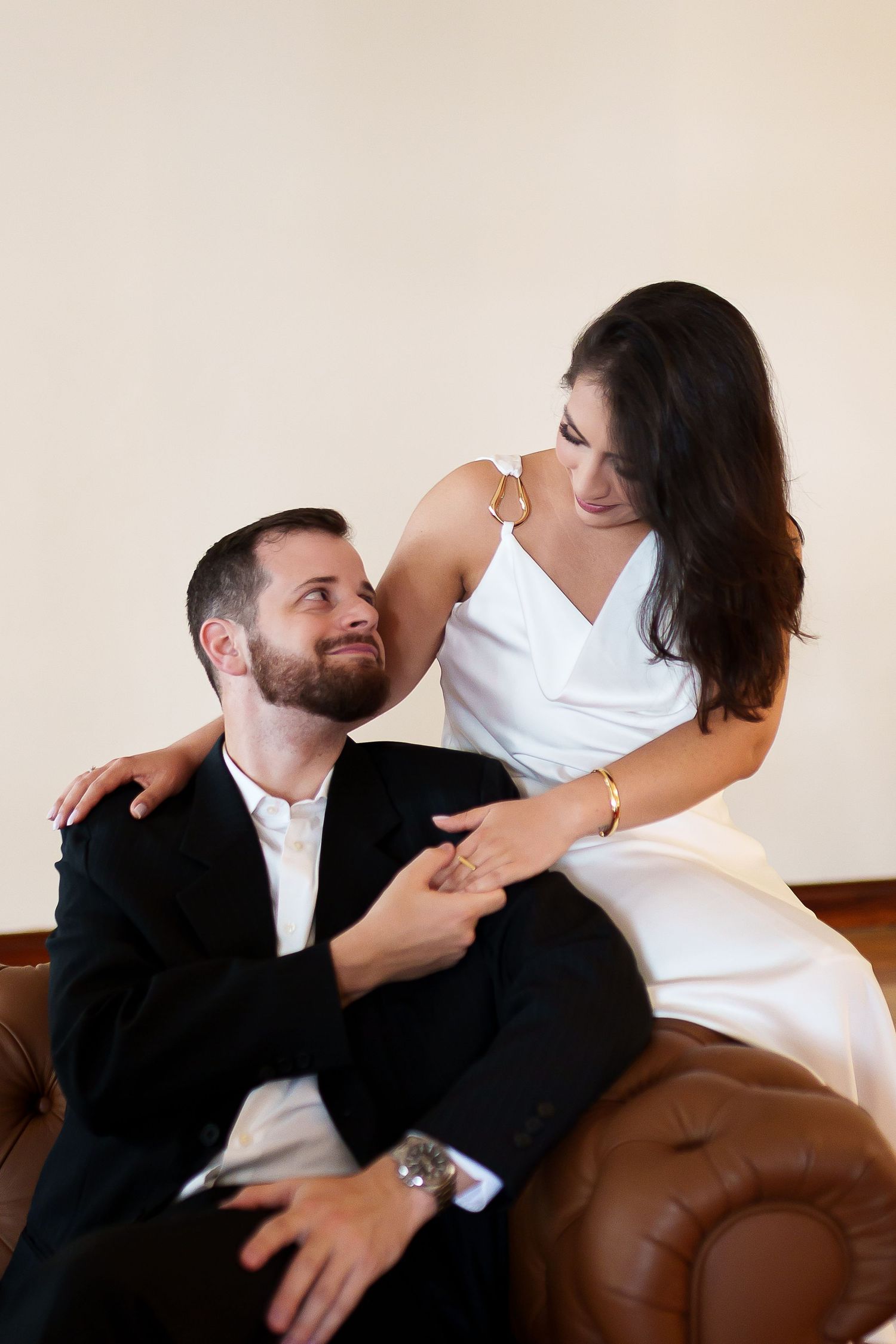 A couple in formal attire sharing a romantic moment on a leather sofa, the woman in white dress sitting beside a bearded man in a suit.
