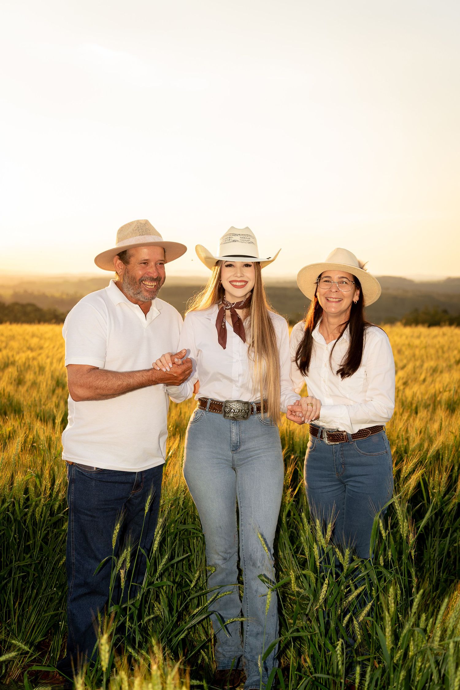 Three people in cowboy hats and white shirts smile in a golden wheat field at sunset.