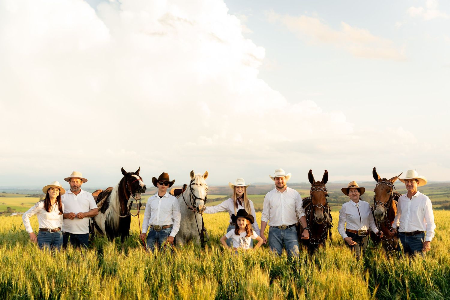 A family in cowboy hats poses with horses in a lush green field under a cloudy sky.