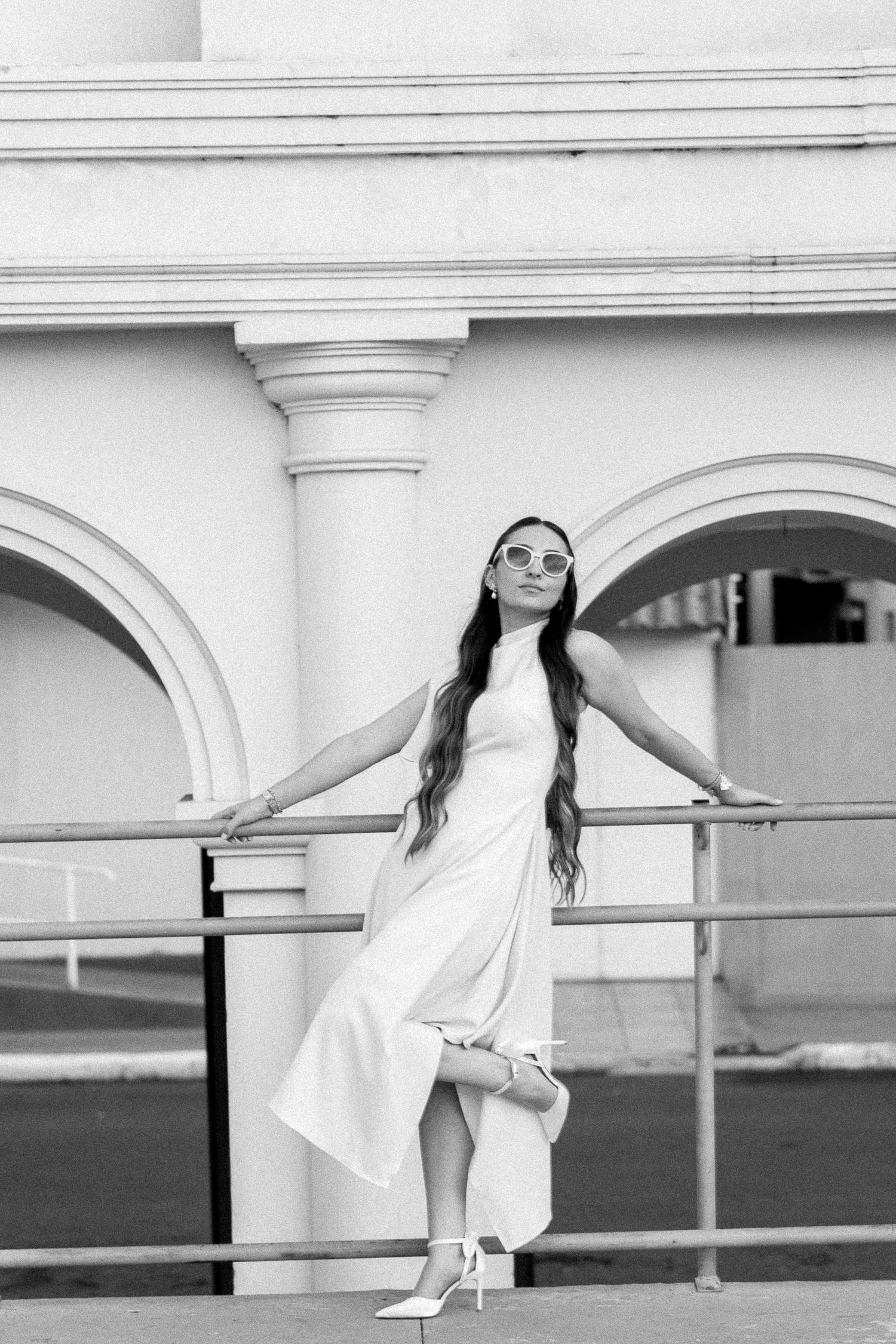 Black and white photo of elegant woman in white dress and sunglasses posing by railing near classical arched building.