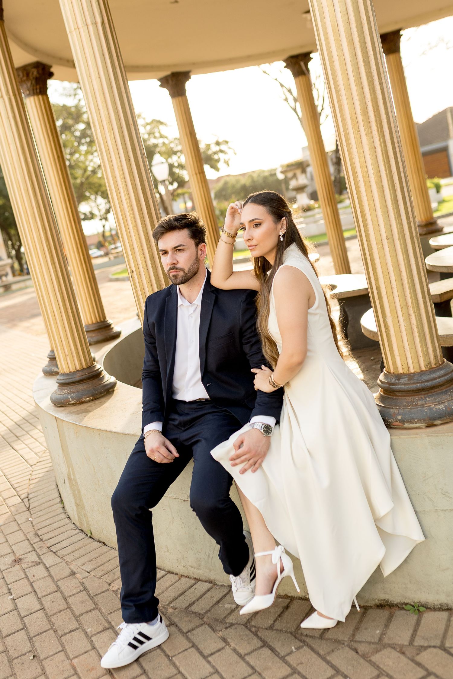 Elegant couple in formal attire posing under classical columns in a park, bride in white dress and groom in dark suit.