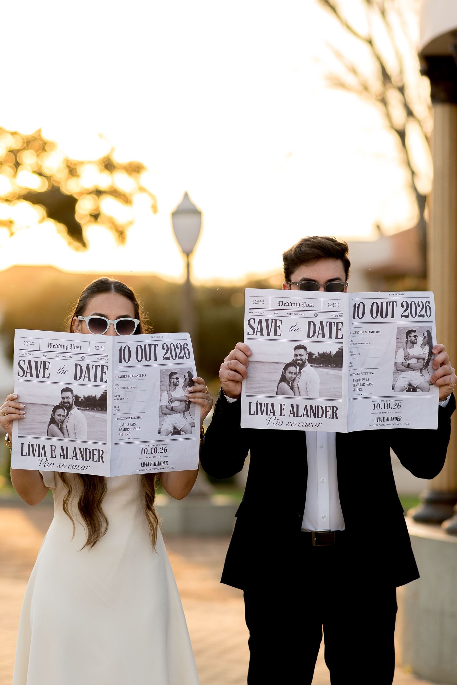 Couple holding Save the Date newspaper announcements for Livia and Alander's October 2026 wedding outdoors at golden hour.