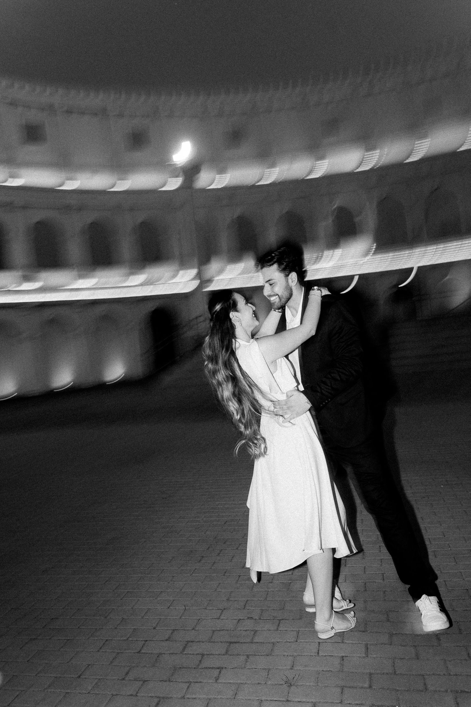 Black and white photo of a couple dancing romantically at night in front of an illuminated historic amphitheater.