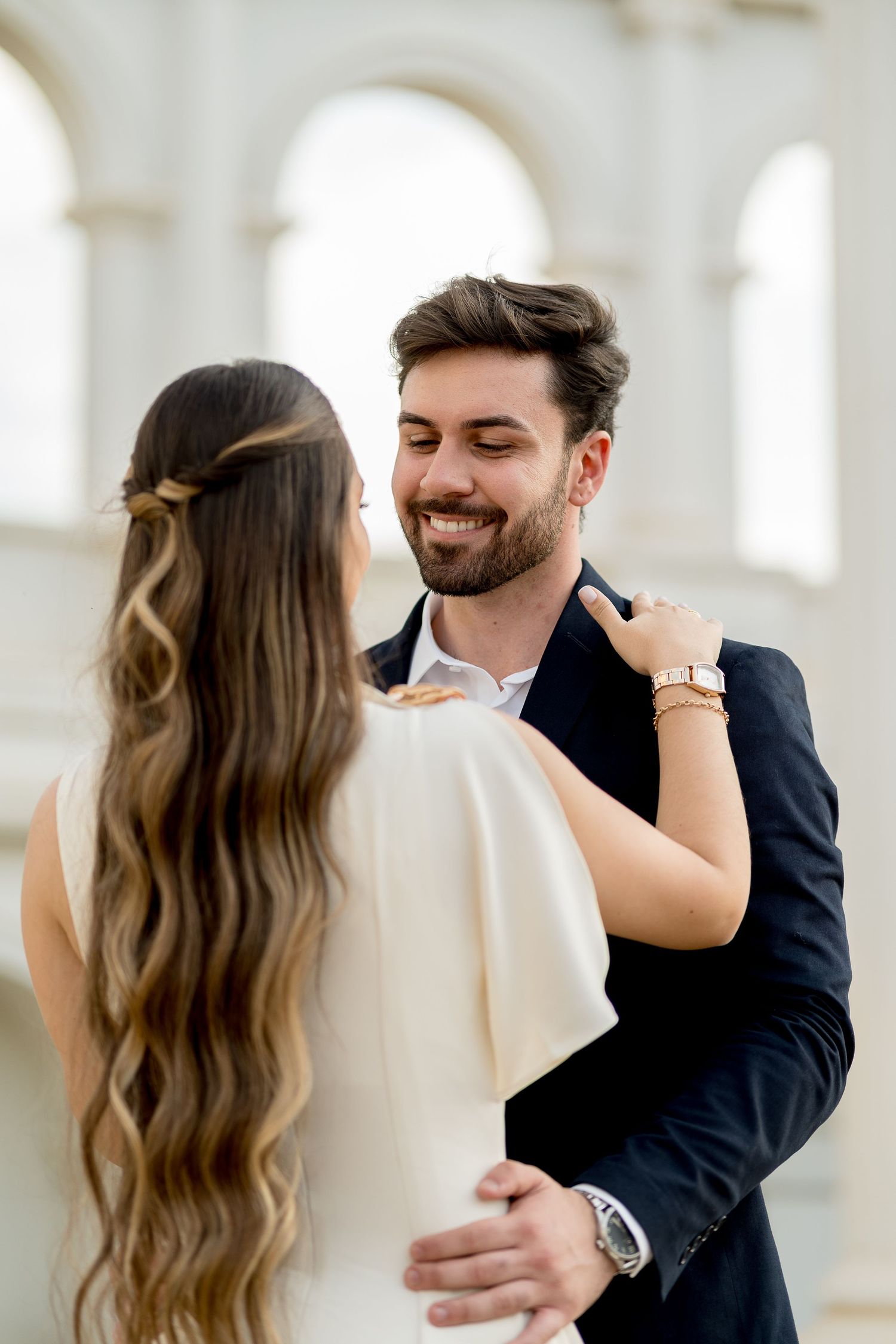 A smiling groom in a navy suit embraces his bride in a white dress, sharing a romantic moment at an elegant venue.