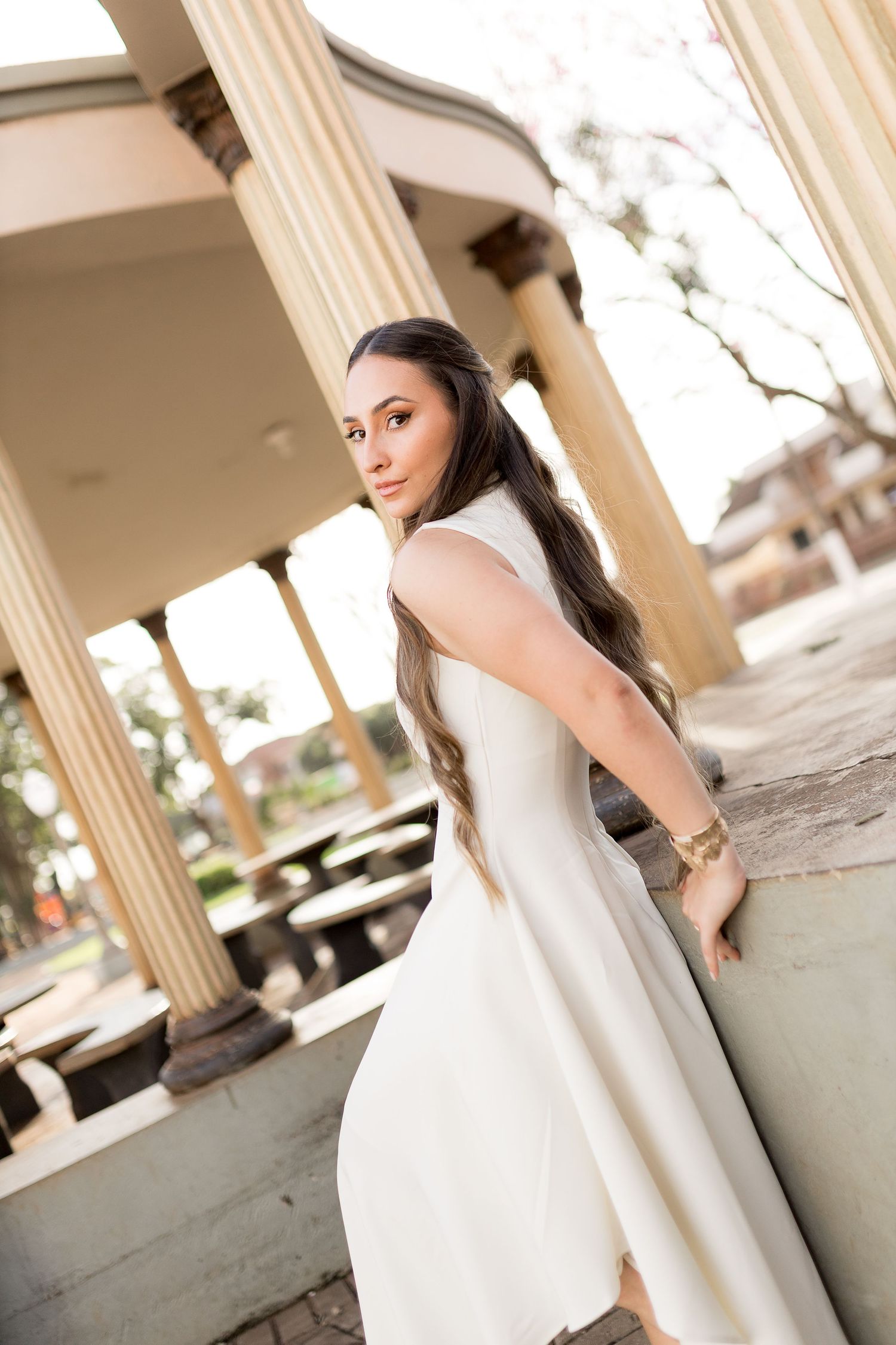 Woman in white dress posing gracefully by classical columns in an outdoor pavilion setting.