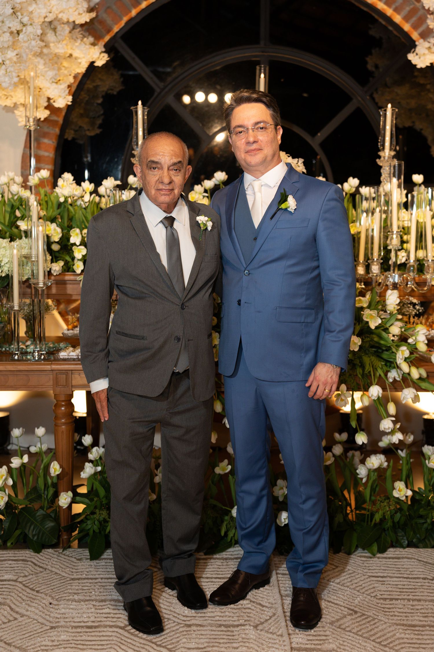 Two men in suits posing together at an elegant wedding reception with white floral decorations.