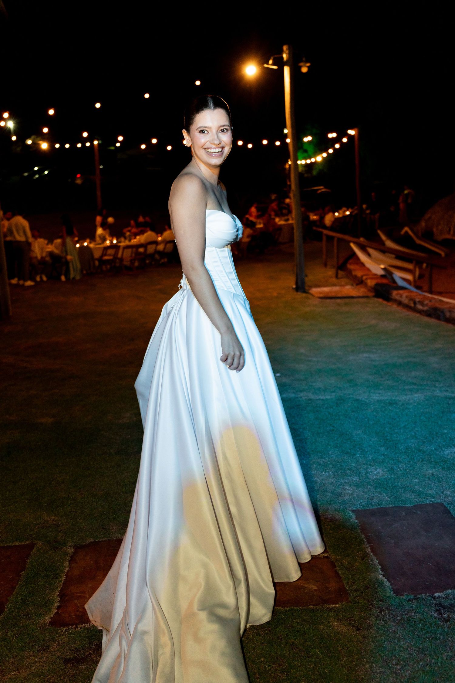 A smiling bride in a flowing white satin gown poses outdoors at night with string lights glowing in the background.