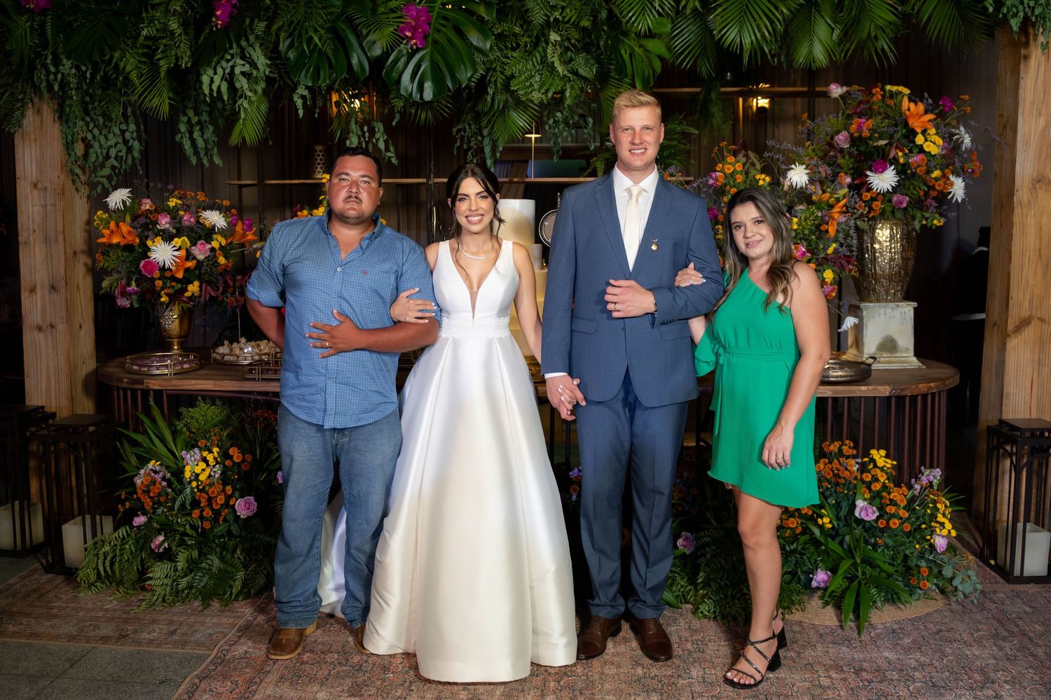 Bride in white gown and groom in blue suit pose with two guests amid lush floral greenery backdrop at wedding reception.