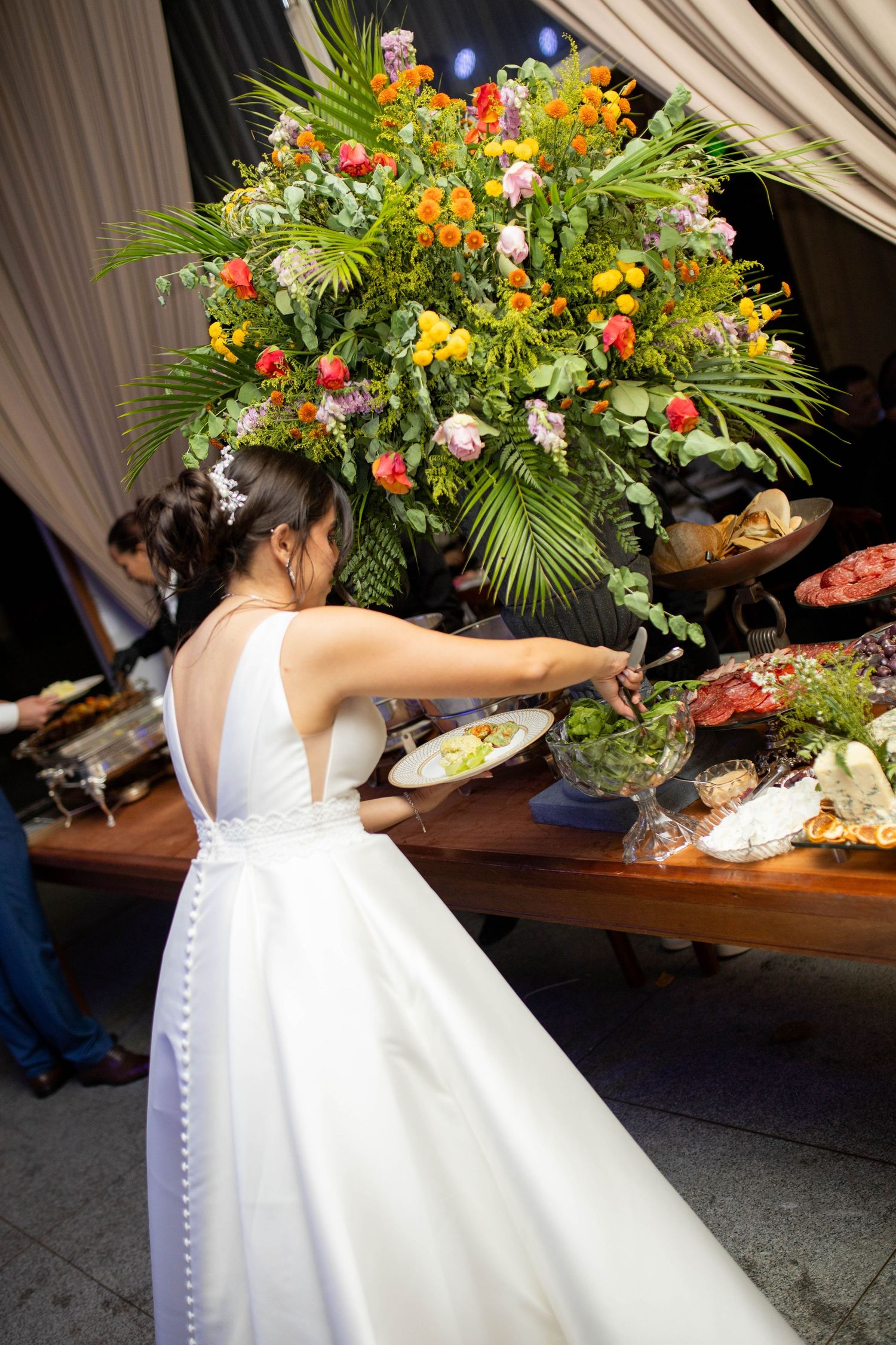 Bride in white gown serving food at an elegant wedding reception buffet table adorned with colorful floral arrangement.