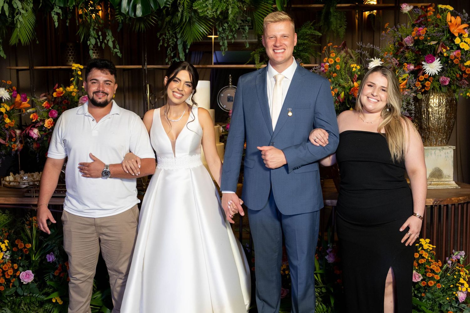 Wedding couple in formal attire poses with two guests amid colorful floral backdrop at elegant reception venue.