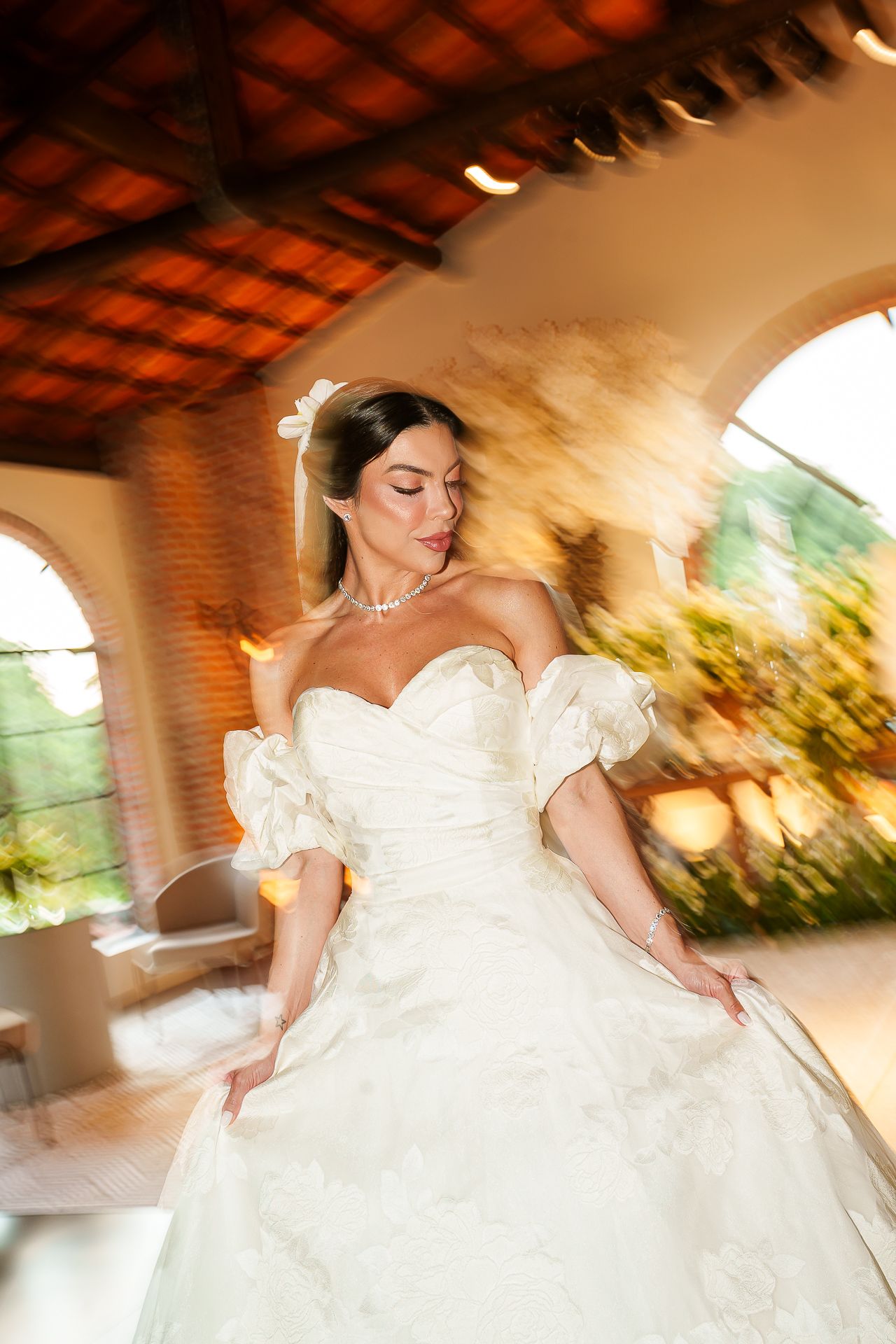 A bride in a white off-shoulder ball gown twirls elegantly in a sunlit venue with arched windows.