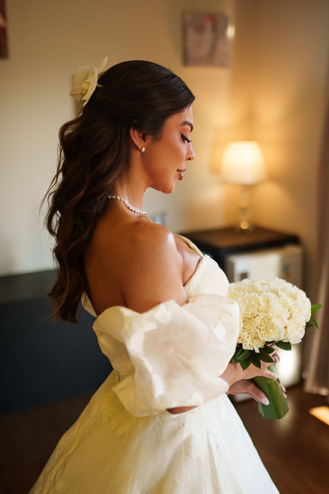 Elegant bride in off-shoulder white gown holding ivory hydrangea bouquet, pearl necklace, soft curls, warm indoor lighting.