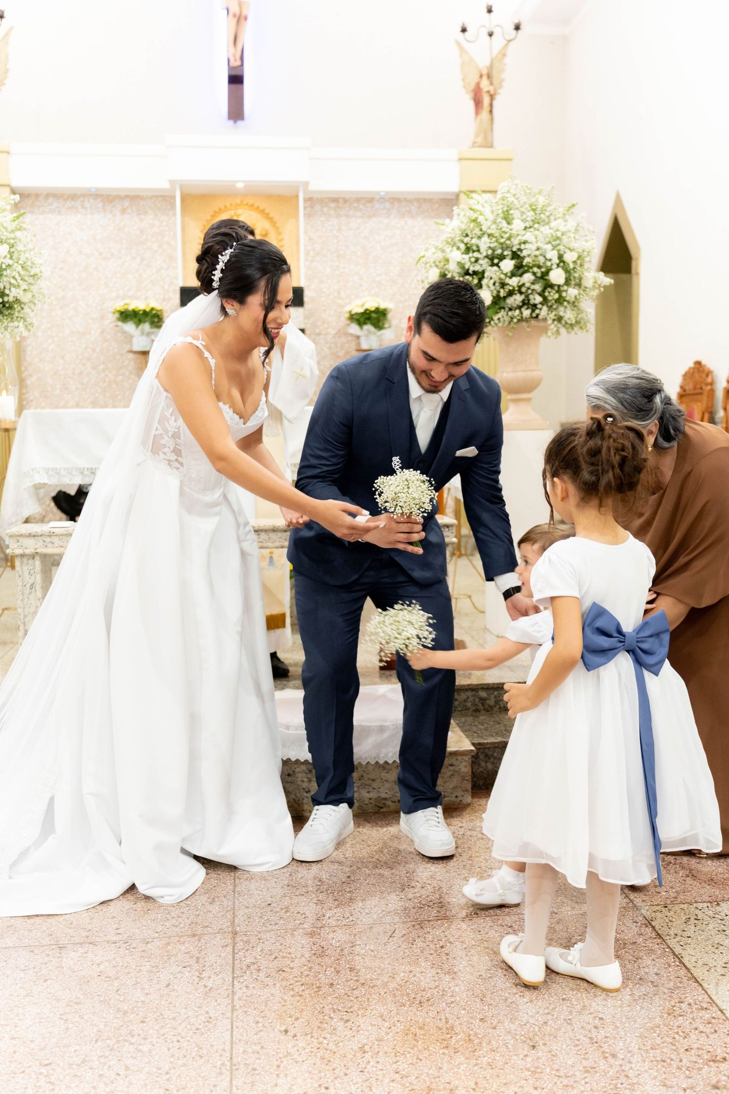 Bride and groom kneel to give flowers to a flower girl in a white dress with blue bow during church wedding ceremony.