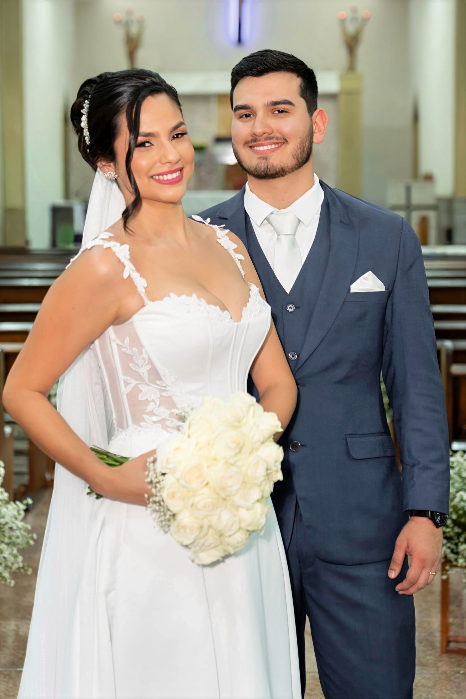 A smiling bride in a white lace gown holding white roses stands beside a groom in a navy suit inside a church.