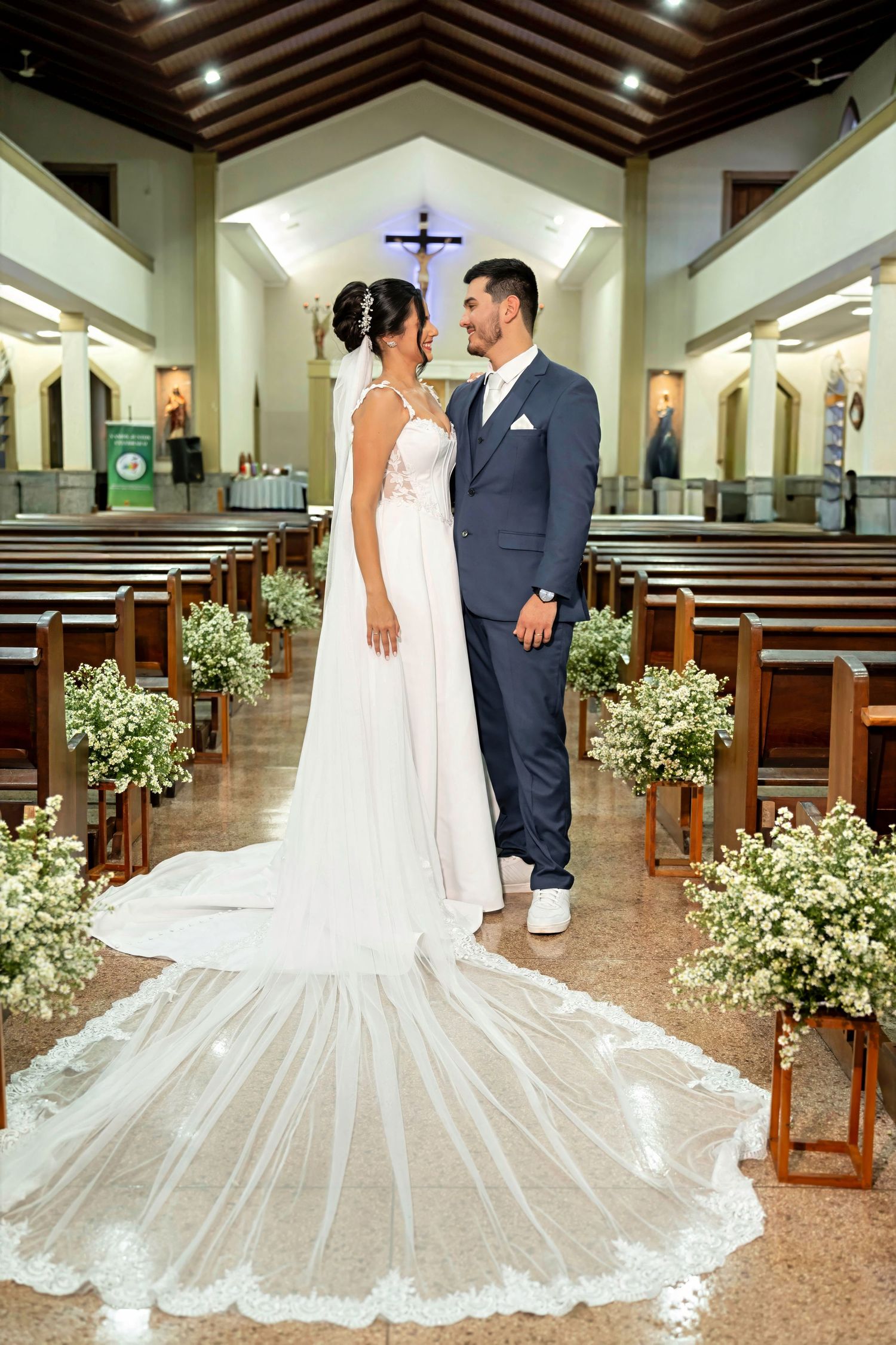 Bride in white gown with long train and groom in navy suit stand together at church altar aisle.