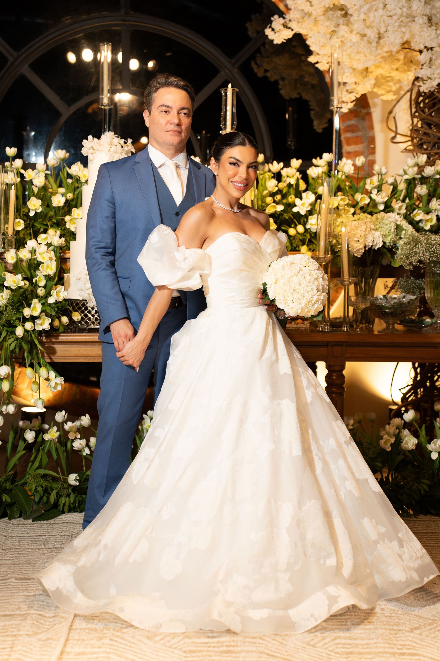 Elegant bride and groom posing together at their wedding reception surrounded by lush white floral arrangements.