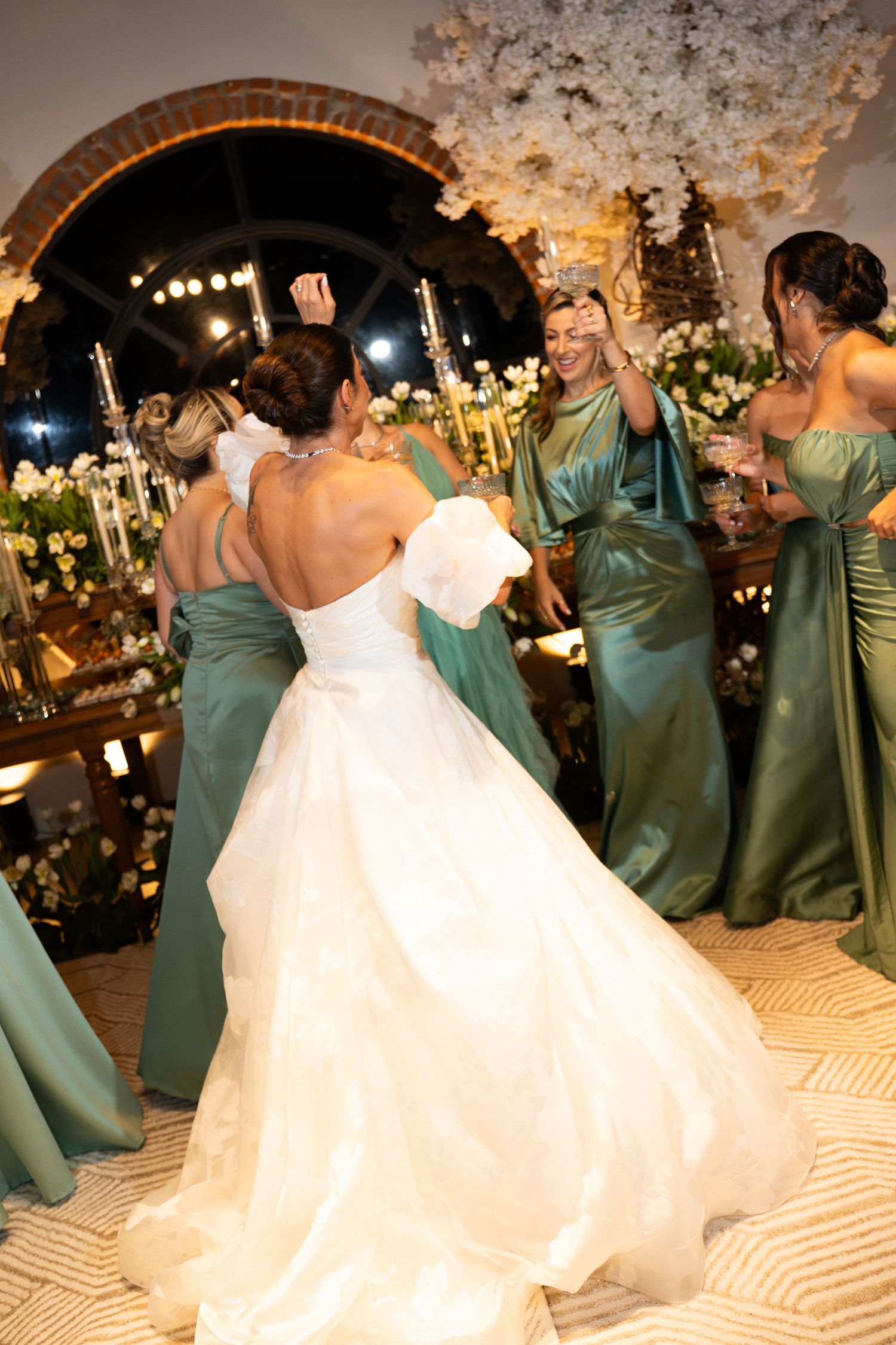 Bride in white gown dancing with bridesmaids in green satin dresses at elegant wedding reception with floral decor.