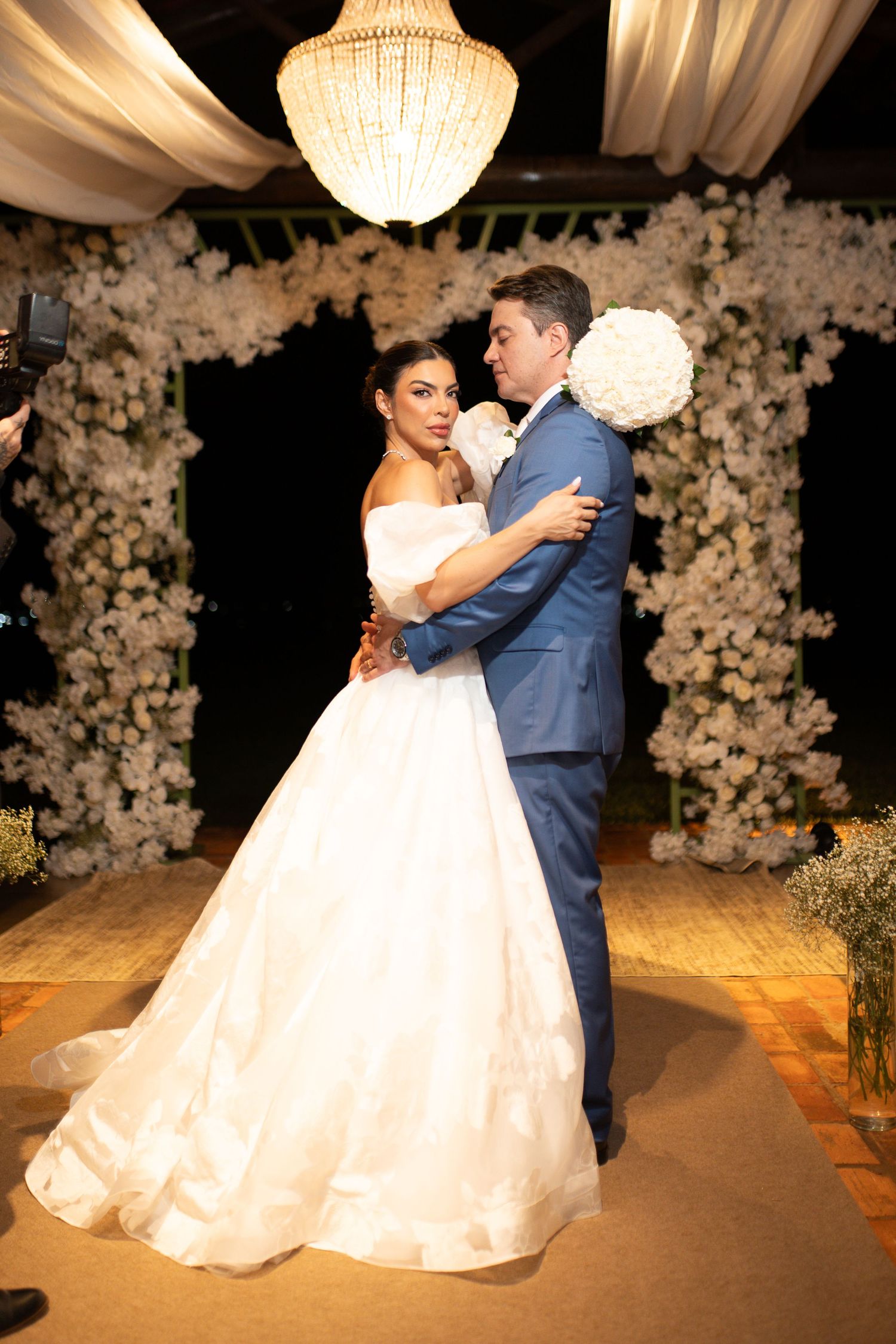 Bride in white ball gown and groom in blue suit share first dance under chandelier with floral arch backdrop.