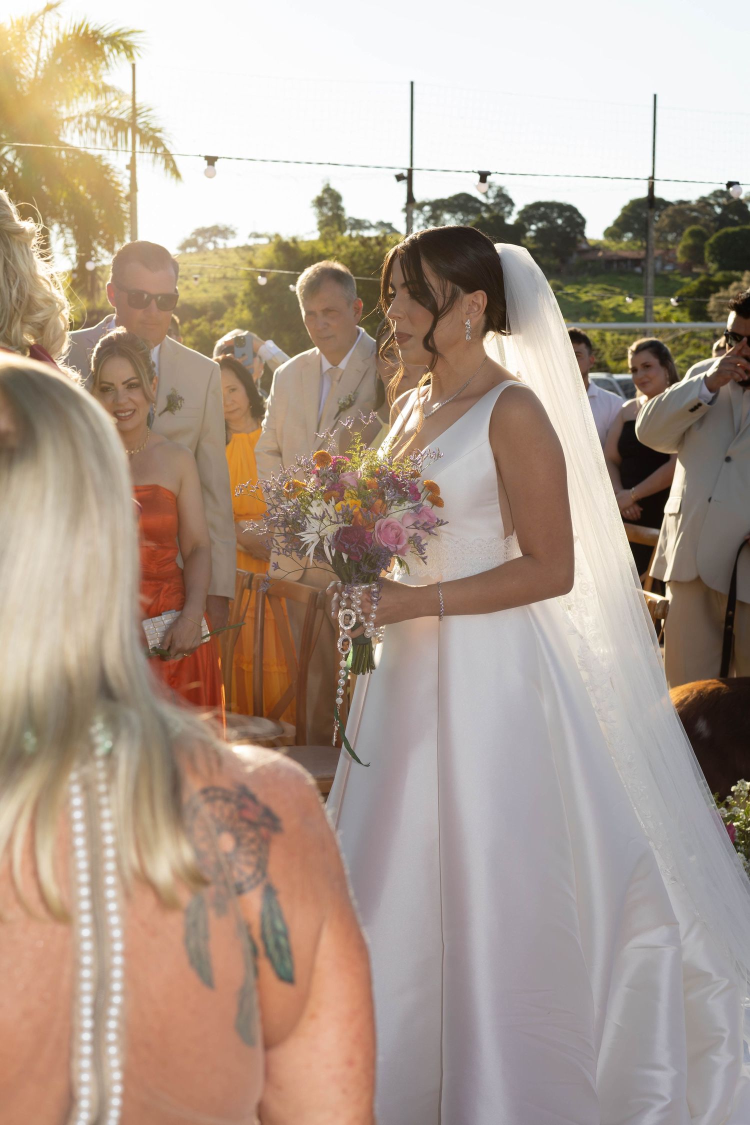 Bride in white gown holding colorful bouquet walks down outdoor aisle, guests watching in golden sunset light.