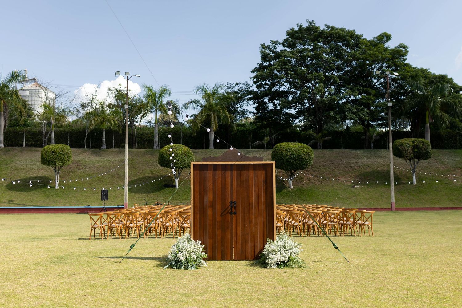 Outdoor wedding ceremony setup on green lawn with wooden doors altar, rows of chairs, and tropical palm trees backdrop.