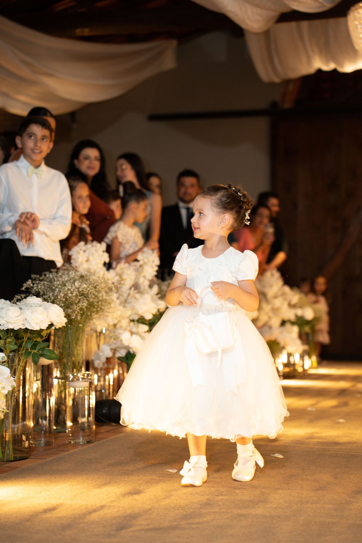 Adorable flower girl in white dress with bow walks down wedding aisle lined with white floral arrangements and candles.