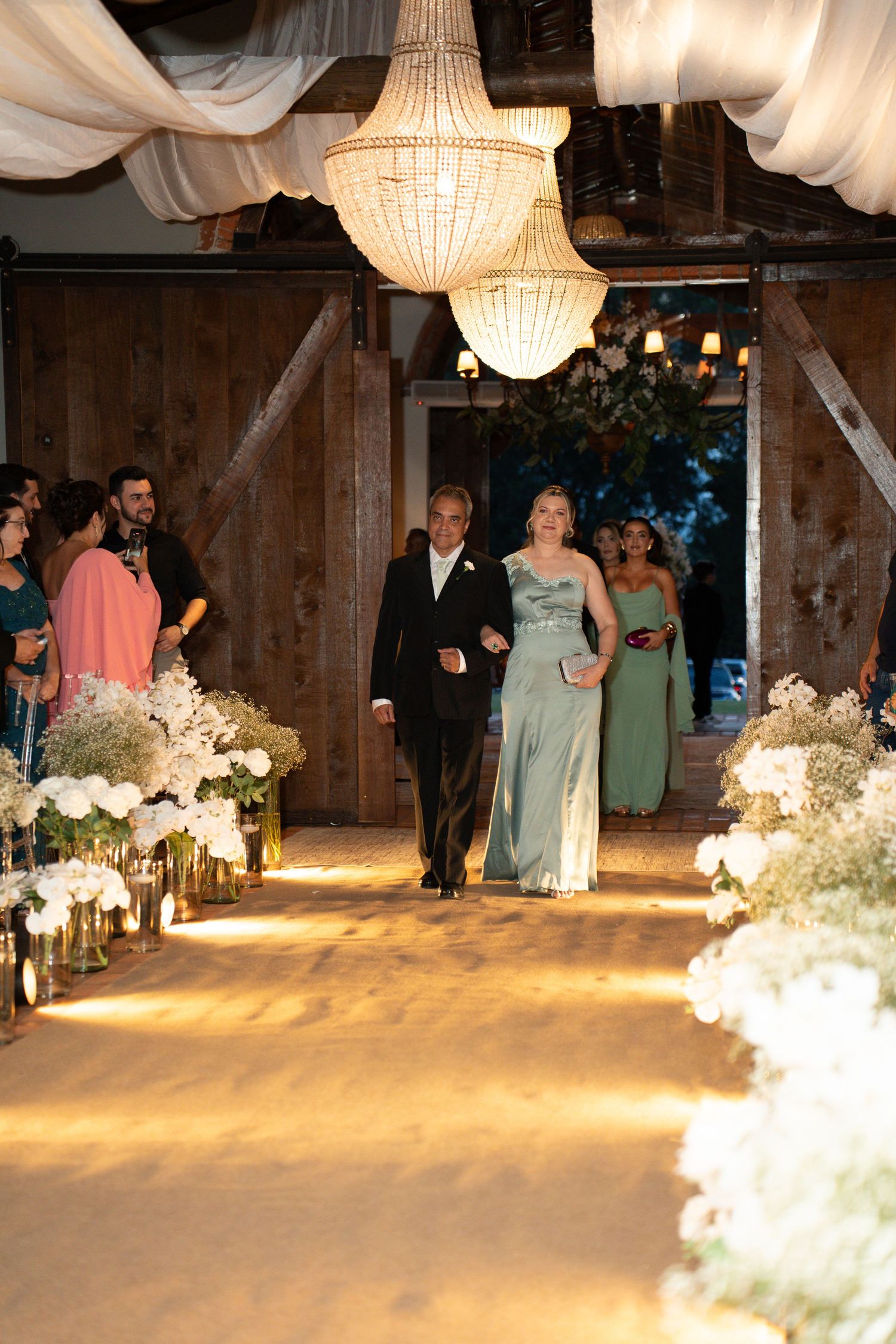 Elegant wedding processional in a barn venue with chandeliers, floral arrangements, and guests watching a couple walk the aisle.