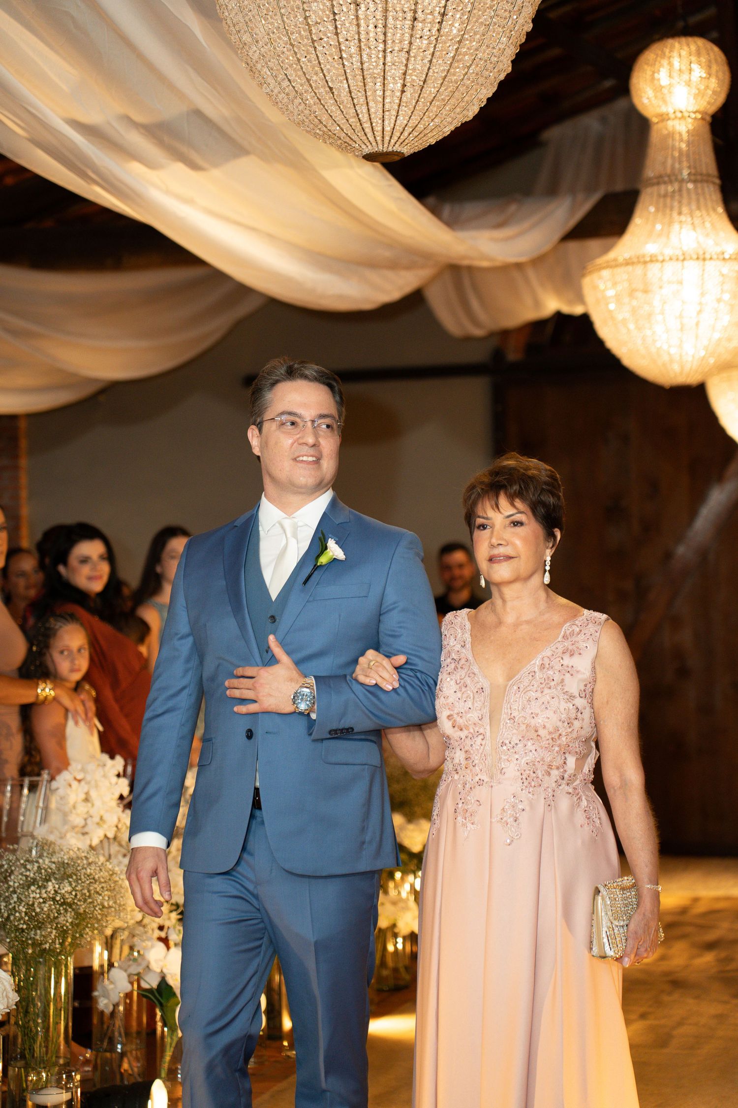 Groom in blue suit escorts elegantly dressed older woman down wedding aisle under crystal chandeliers.