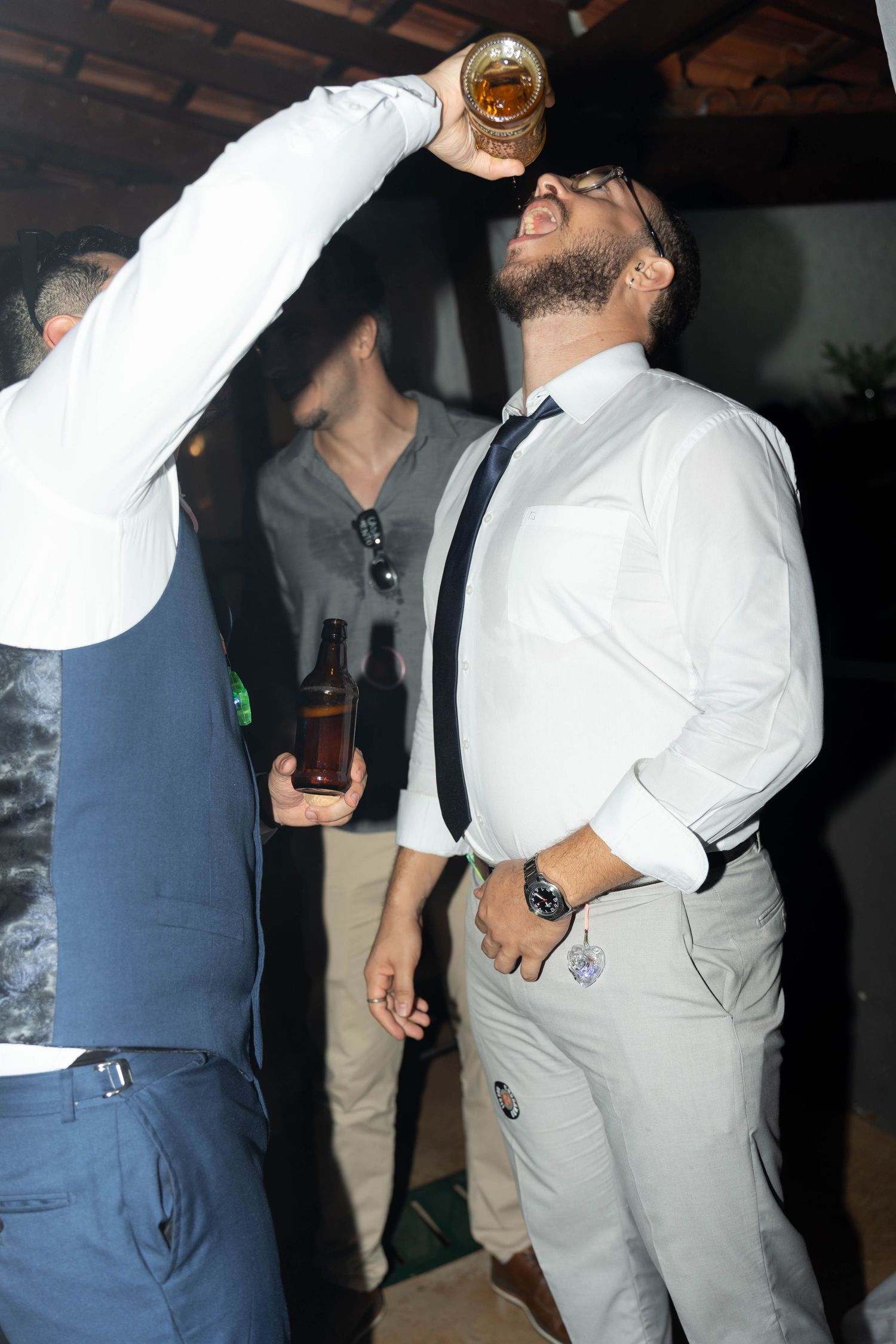 Man in white shirt and tie drinking from raised glass at a party with friends holding beer bottles.