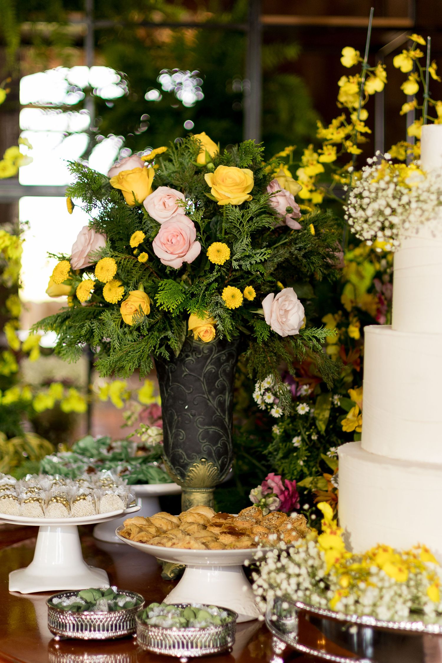 Elegant wedding dessert table with white tiered cake, yellow and pink roses in dark vase, and assorted sweets.