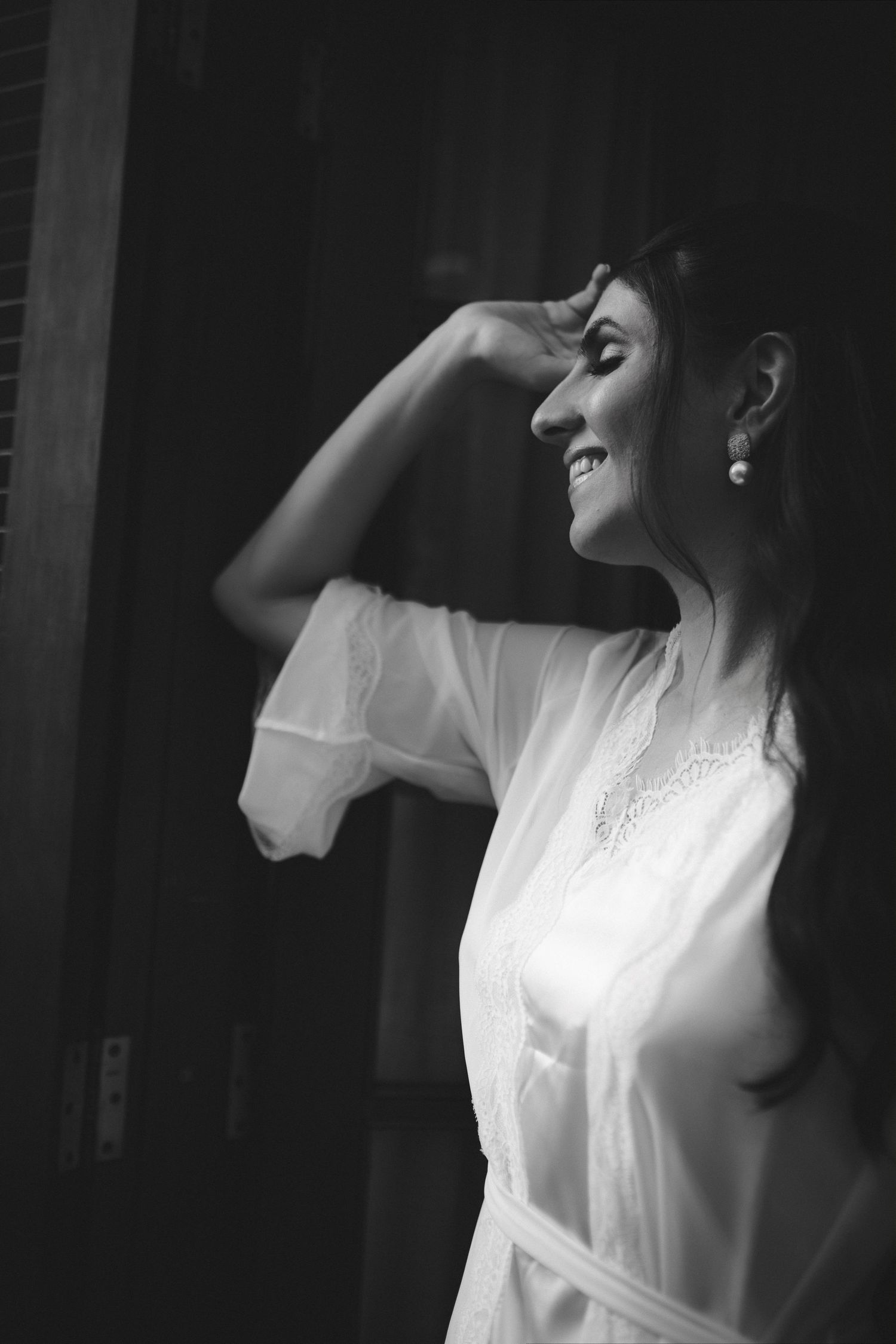 Black and white photo of a smiling bride in a white robe, hand in hair, standing in soft light.