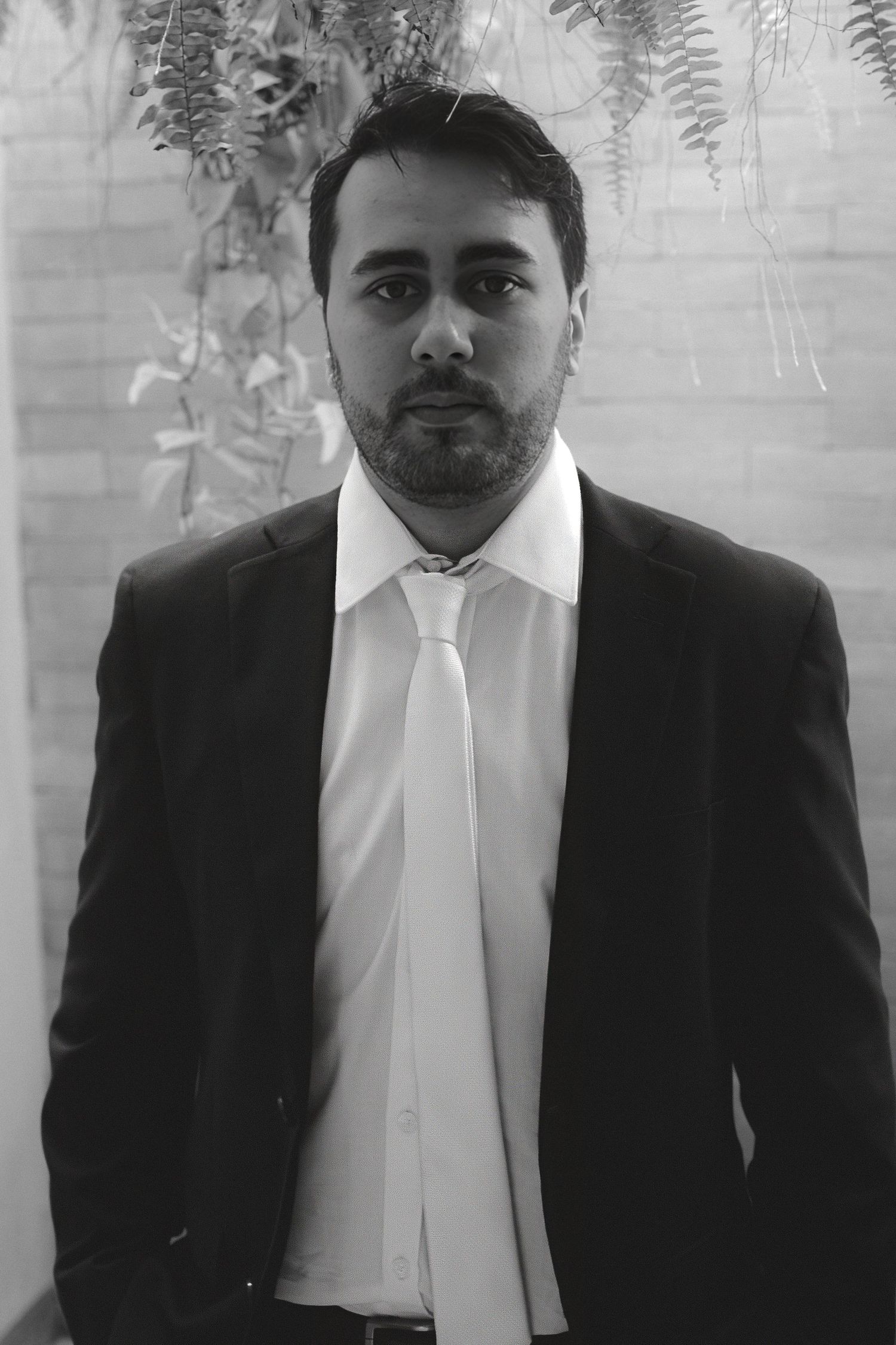 Black and white portrait of a bearded man in a suit and tie standing before lush green foliage.