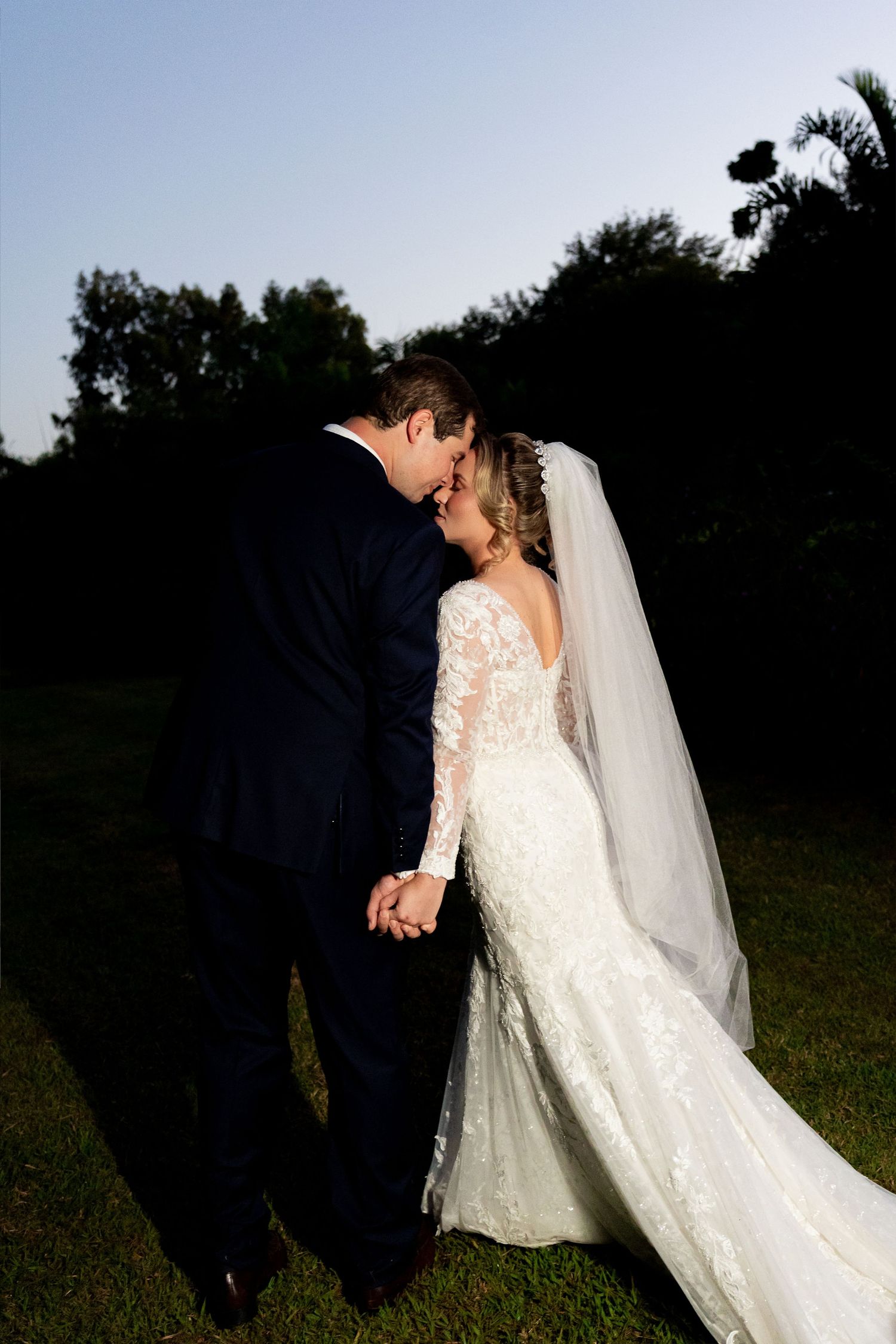 Bride in lace gown and veil holds hands with groom in navy suit, sharing a romantic moment outdoors at dusk.